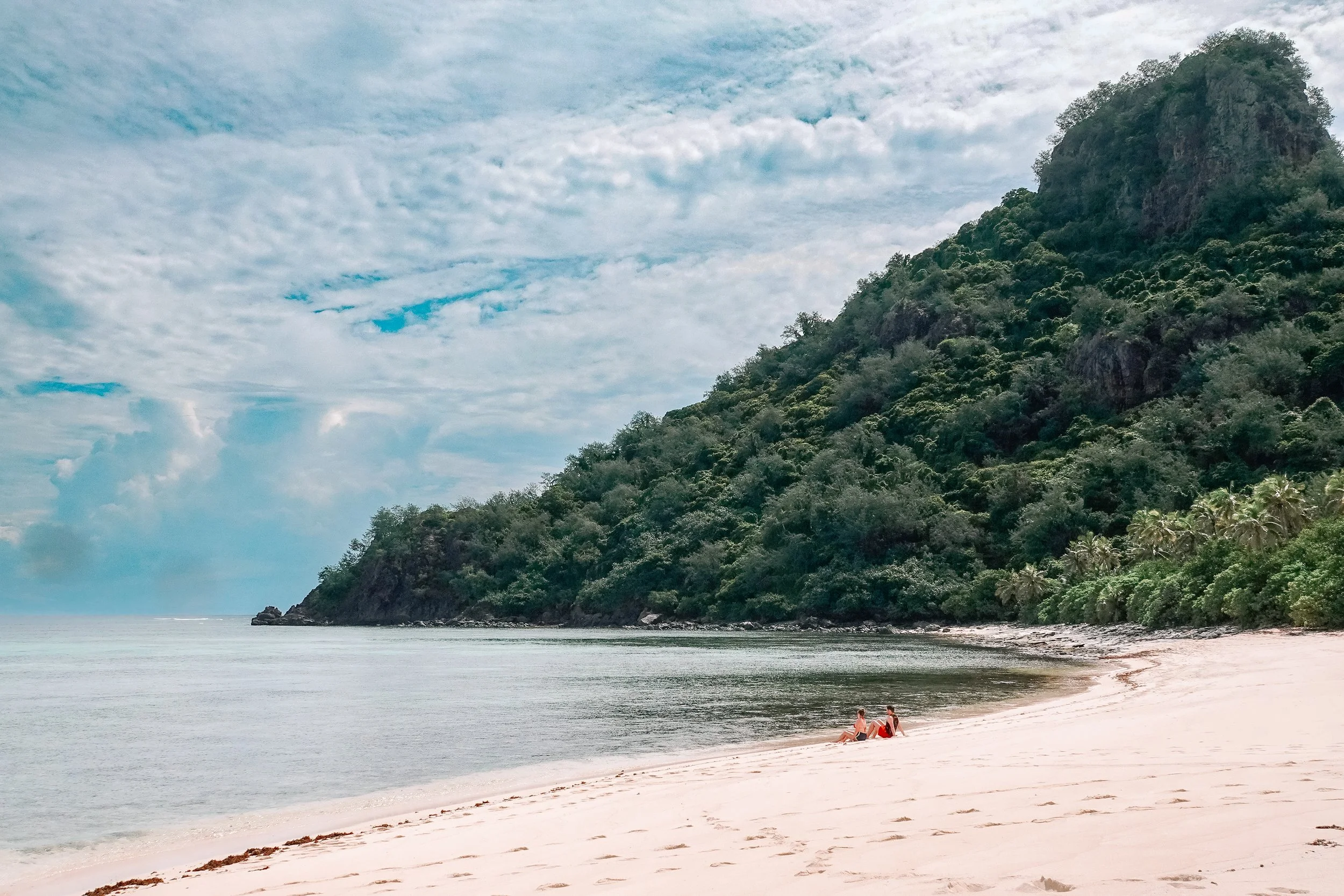 A tranquil beach scene with two women sitting on the sandy shore, overlooking calm water and a lush green hillside under a partly cloudy sky.