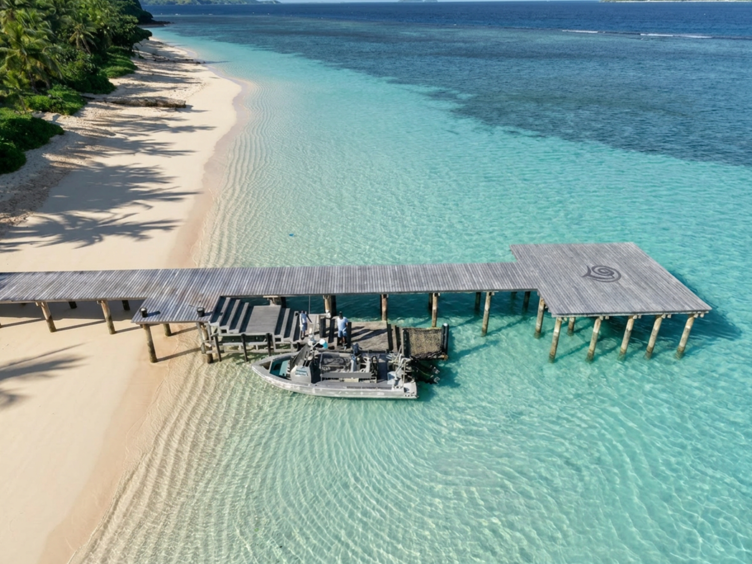 An aerial view of a tropical beach with clear turquoise water, white sandy shore, lush green trees, and a dock with a boat.