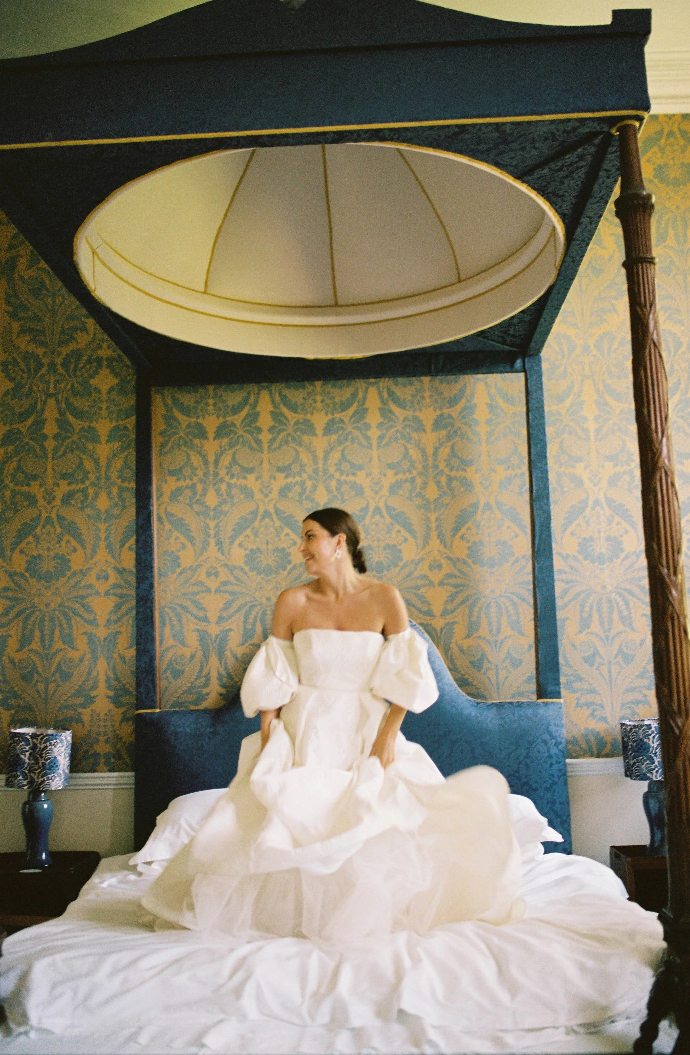 A woman in a white off-the-shoulder dress sits on a bed in a room with ornate wallpaper. She is smiling and looking to her left. Above the bed is a large canopy or frame with dark blue fabric and gold trim, and an oval mirror is embedded in the top p