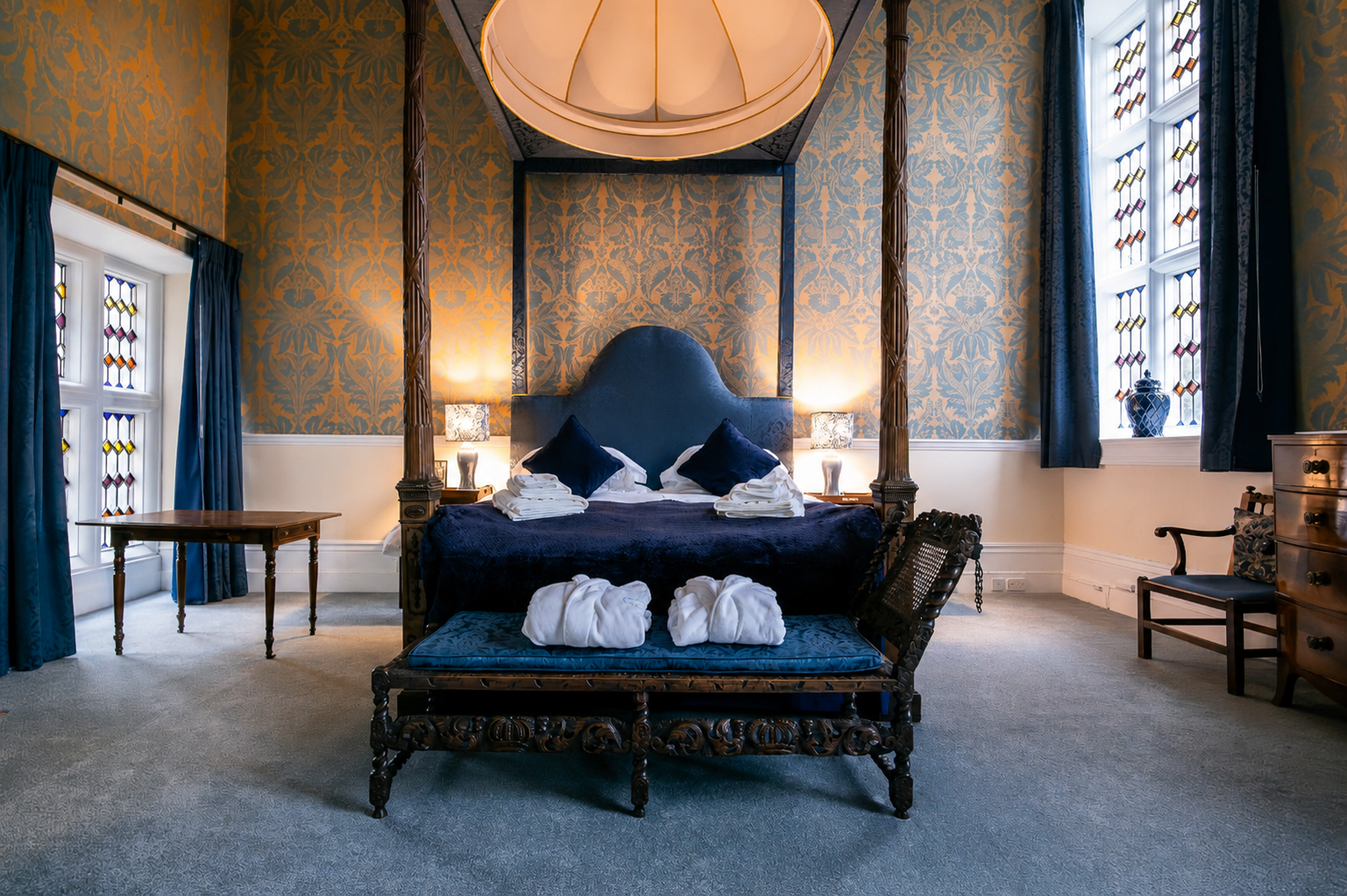 Elegant bedroom with a large four-poster bed with a blue headboard and dark blue bedding, four towels folded on the bed, and two blue pillows. The walls have ornate wallpaper with a gold pattern, and there are two windows with stained glass and blue 