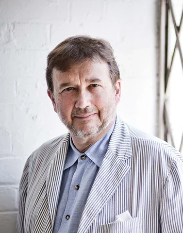 A middle-aged man with brown hair and a beard, wearing a striped suit jacket and a blue shirt, standing indoors near a window with white brick walls in the background.