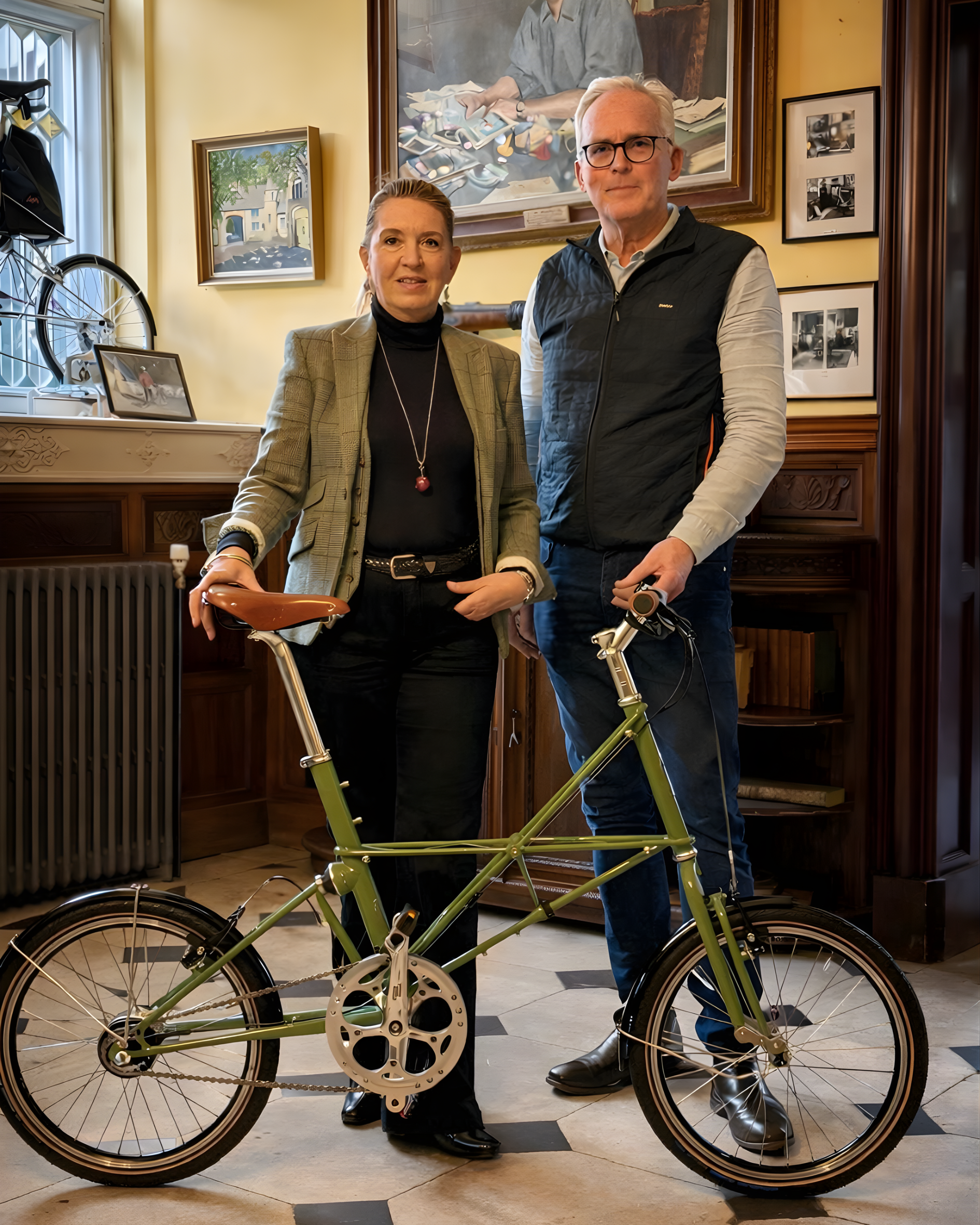 A man and a woman standing indoors with a green folding bicycle. The woman is wearing a blazer, black turtleneck, and black pants, and the man is wearing a light-colored shirt, navy vest, and dark pants. The background features paintings, framed photographs, a window, and wooden furniture.