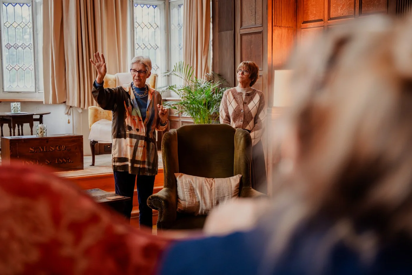 A group of people standing on a lawn in front of a historic building with intricate architectural details, including large windows and ornate stonework. The scene includes greenery, such as trees and bushes, under a clear blue sky.