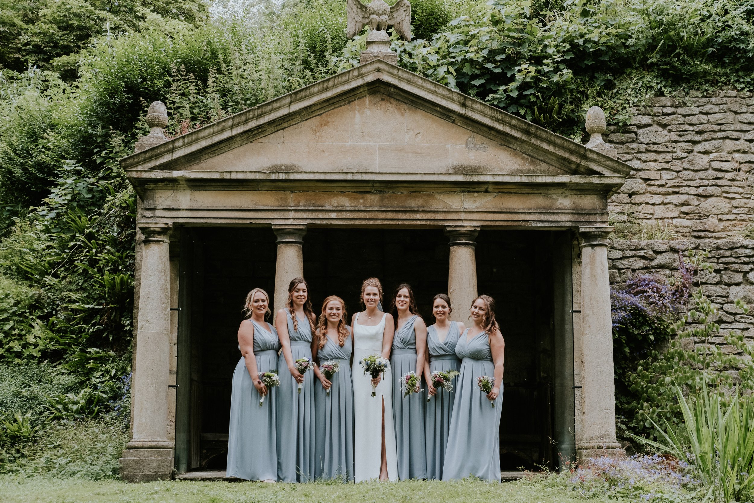 A group of women in matching light blue bridesmaid dresses standing in front of a classical stone structure with columns in a garden setting.