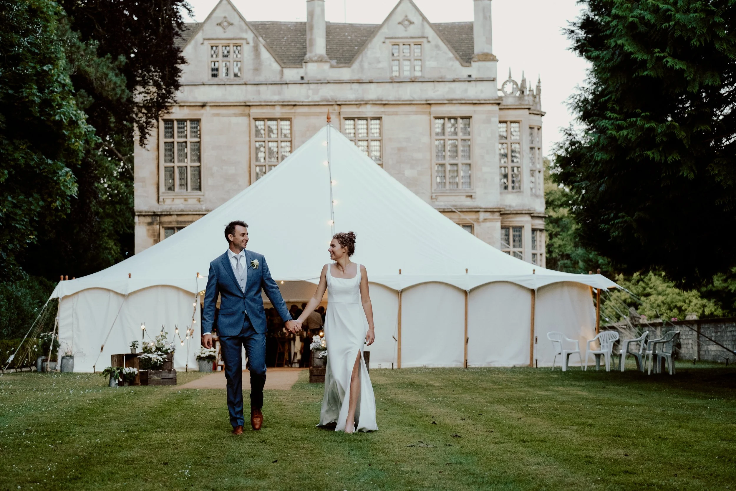 A bride and groom holding hands and walking outside a white wedding tent, with a historic stone building in the background, surrounded by trees and chairs.