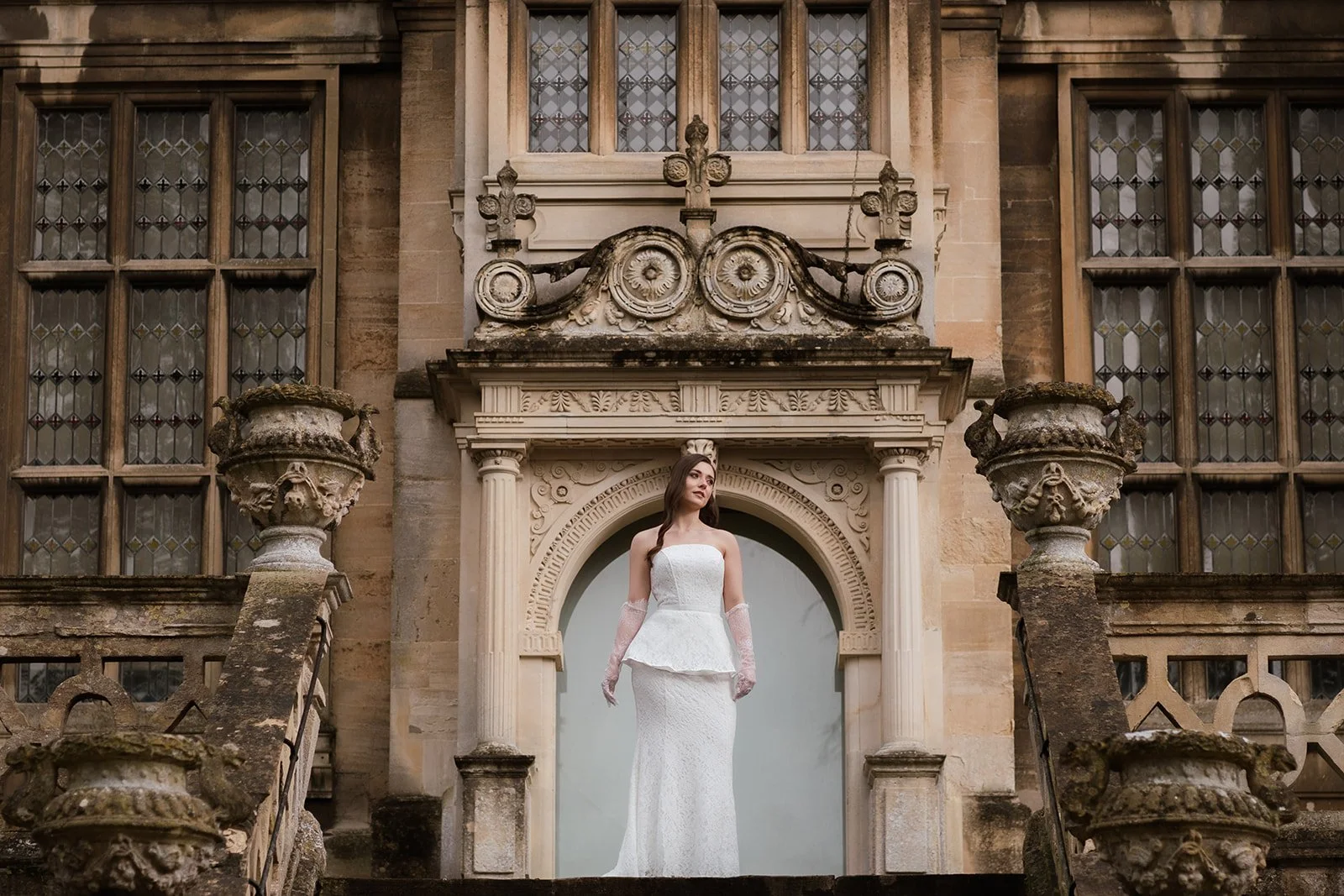 A woman in a white wedding dress standing on the steps of an ornate historic building with large windows and intricate stonework.