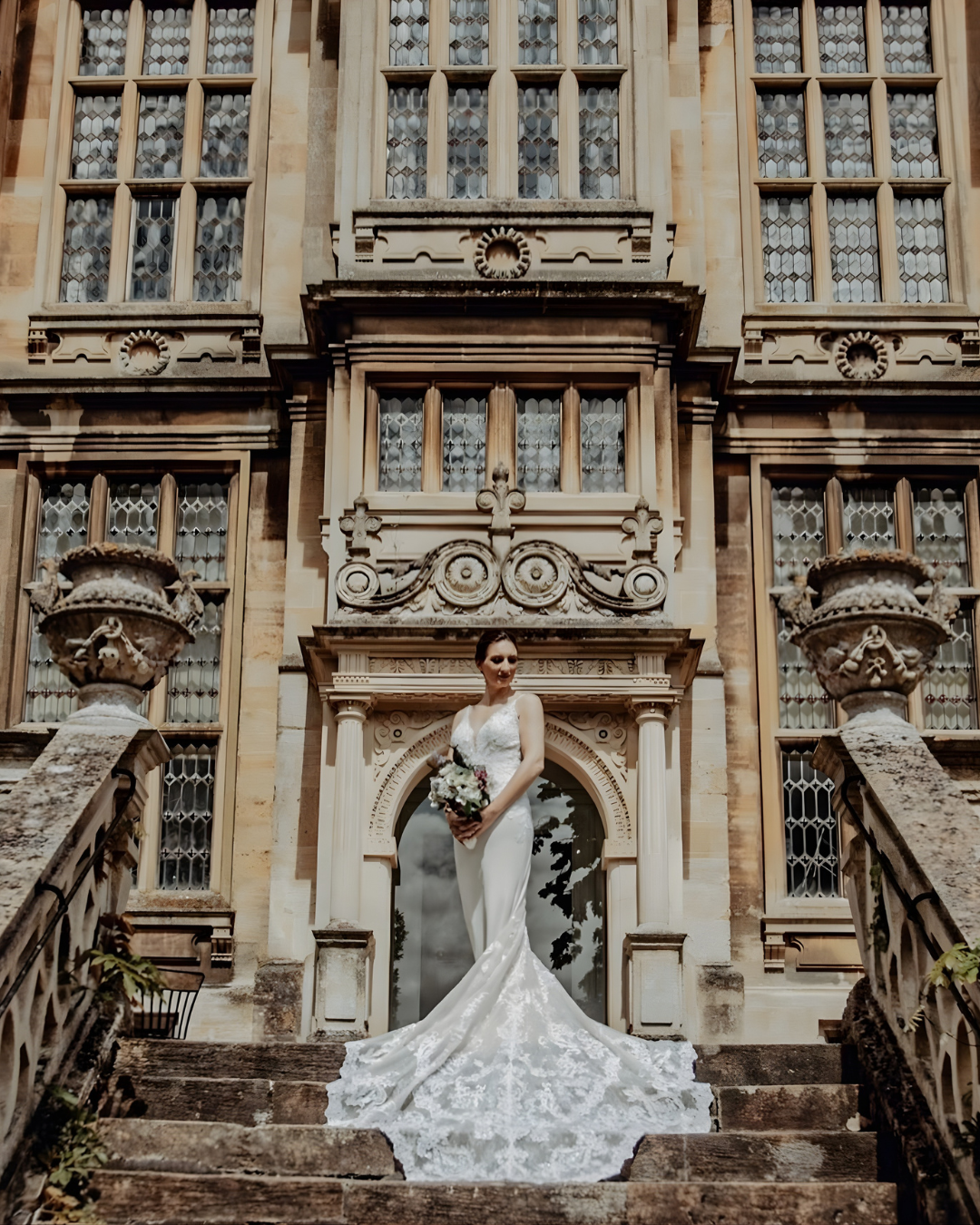 A bride in a white wedding gown holding a bouquet, standing on stone steps in front of a historic building with ornate windows and architectural details.