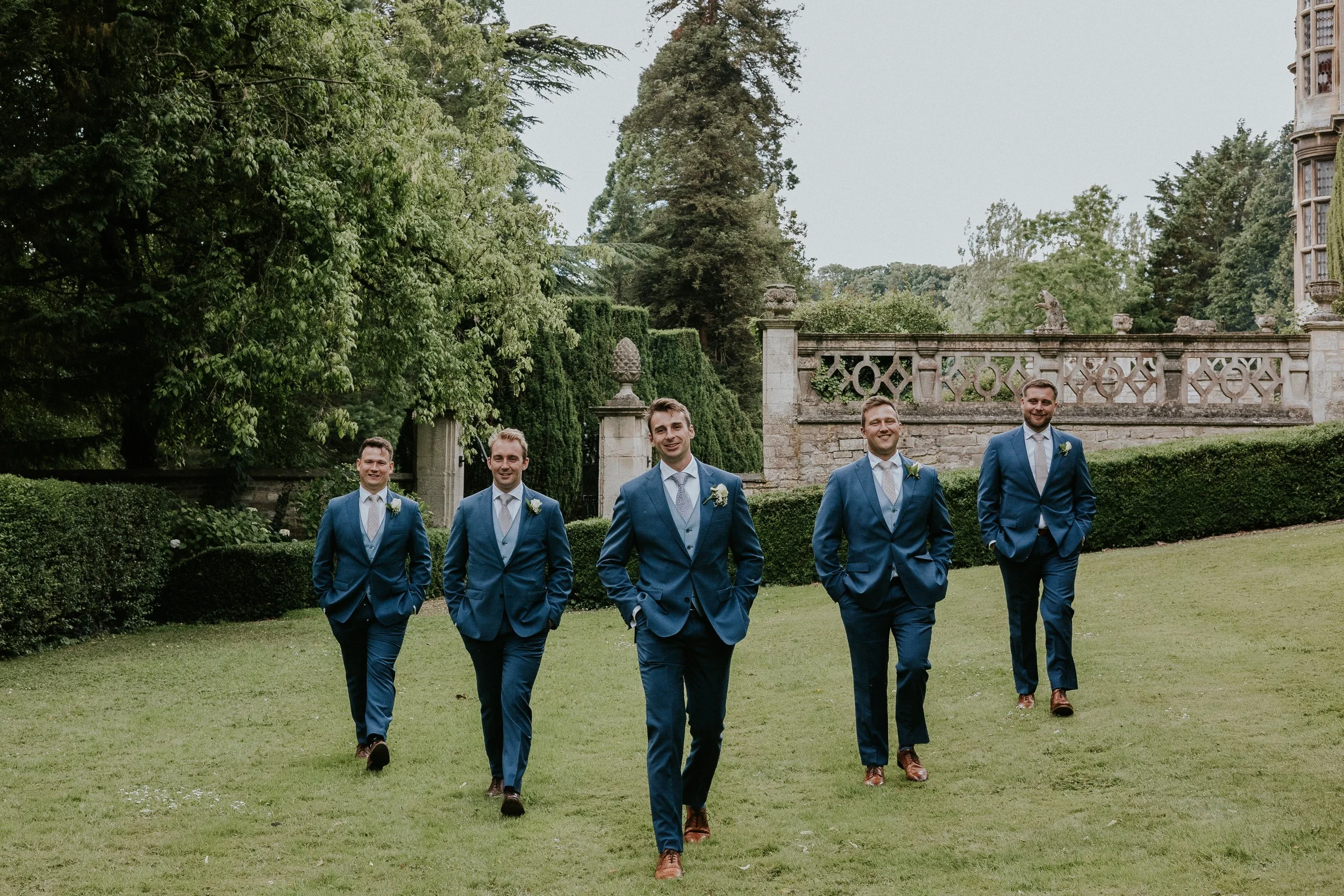 A group of five men dressed in blue suits walking on a grassy lawn outdoors, with a stone wall and trees in the background.