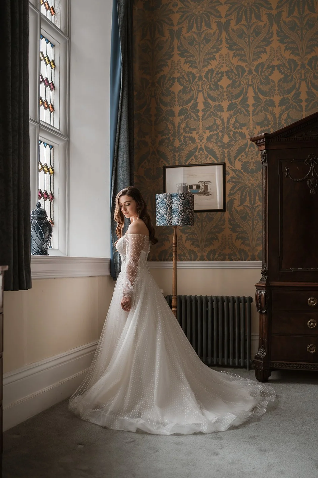 A woman in a white wedding dress standing by a window with stained glass, in a vintage-style room with patterned wallpaper, a framed artwork, and a wooden armoire.