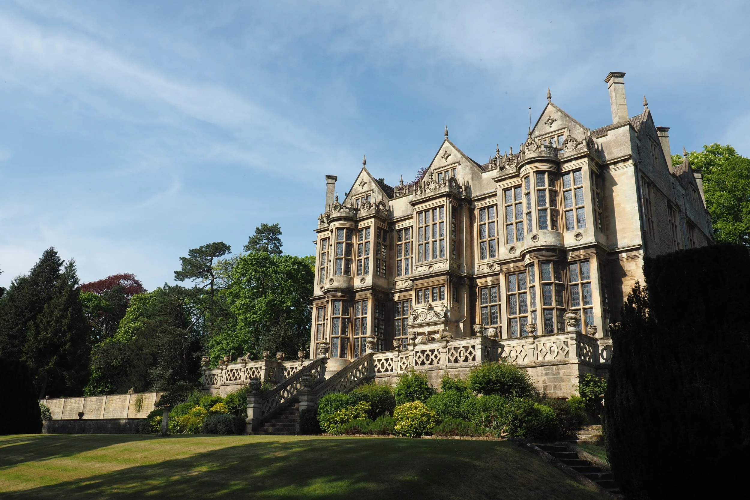 An old stone mansion with tall, intricate windows and decorative stonework, surrounded by lush greenery and trees.