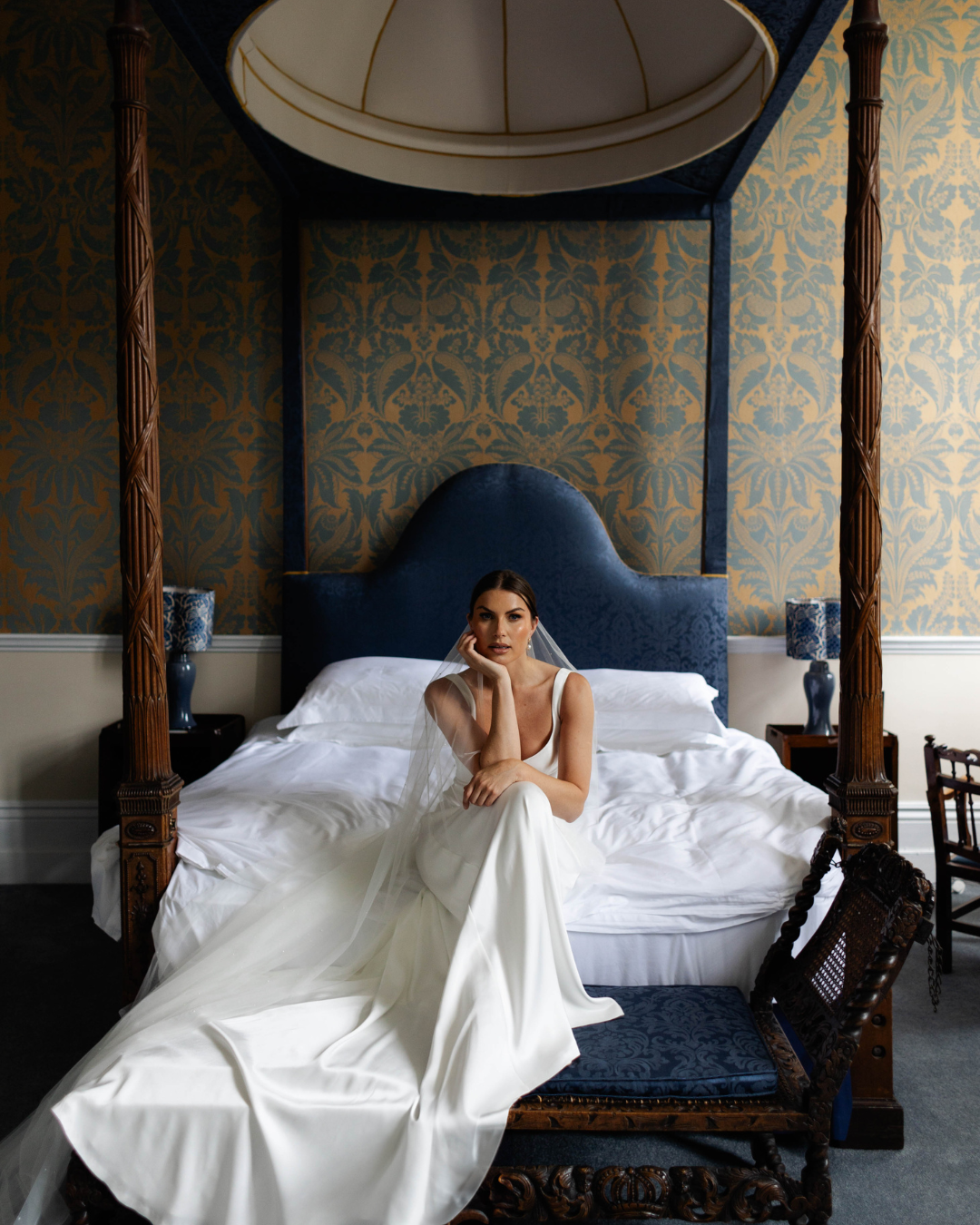 A woman in a white wedding dress with a veil sits on the edge of a large, ornate four-poster bed in a room with elegant gold and blue patterned wallpaper, blue lamps on dark wood side tables.