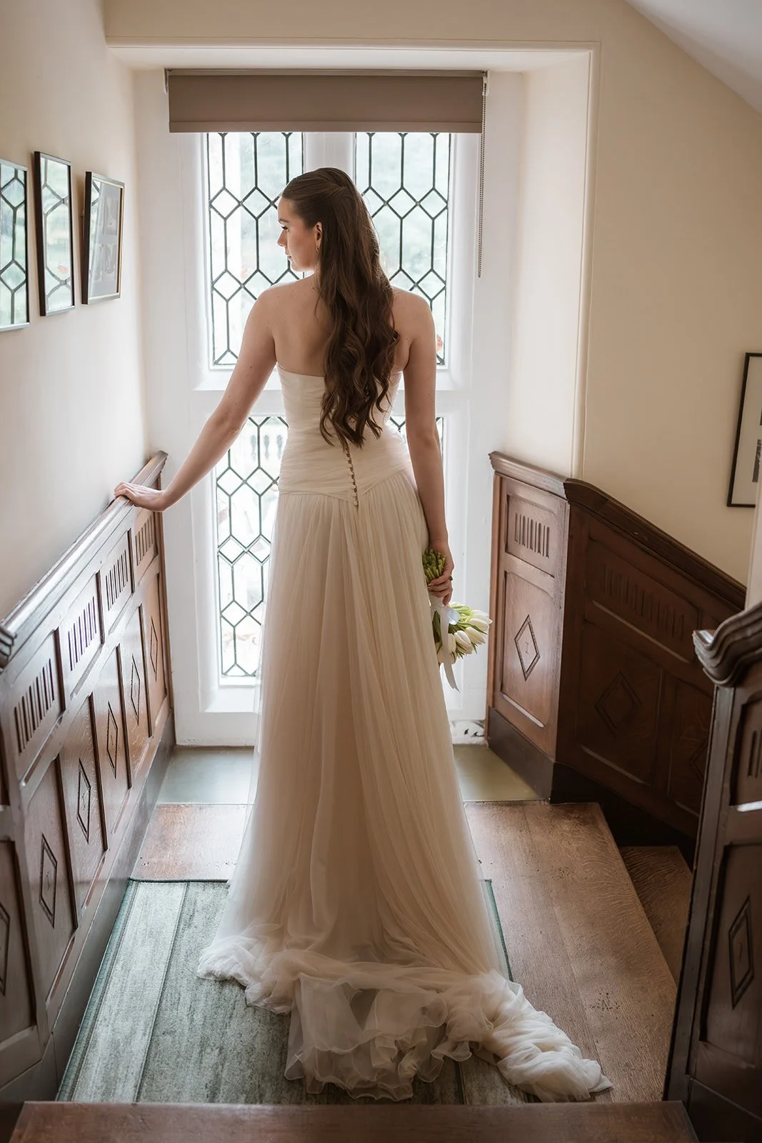 A woman in a strapless wedding dress holding a bouquet of white flowers standing in front of a window with geometric patterned glass, inside a house with wooden paneling.