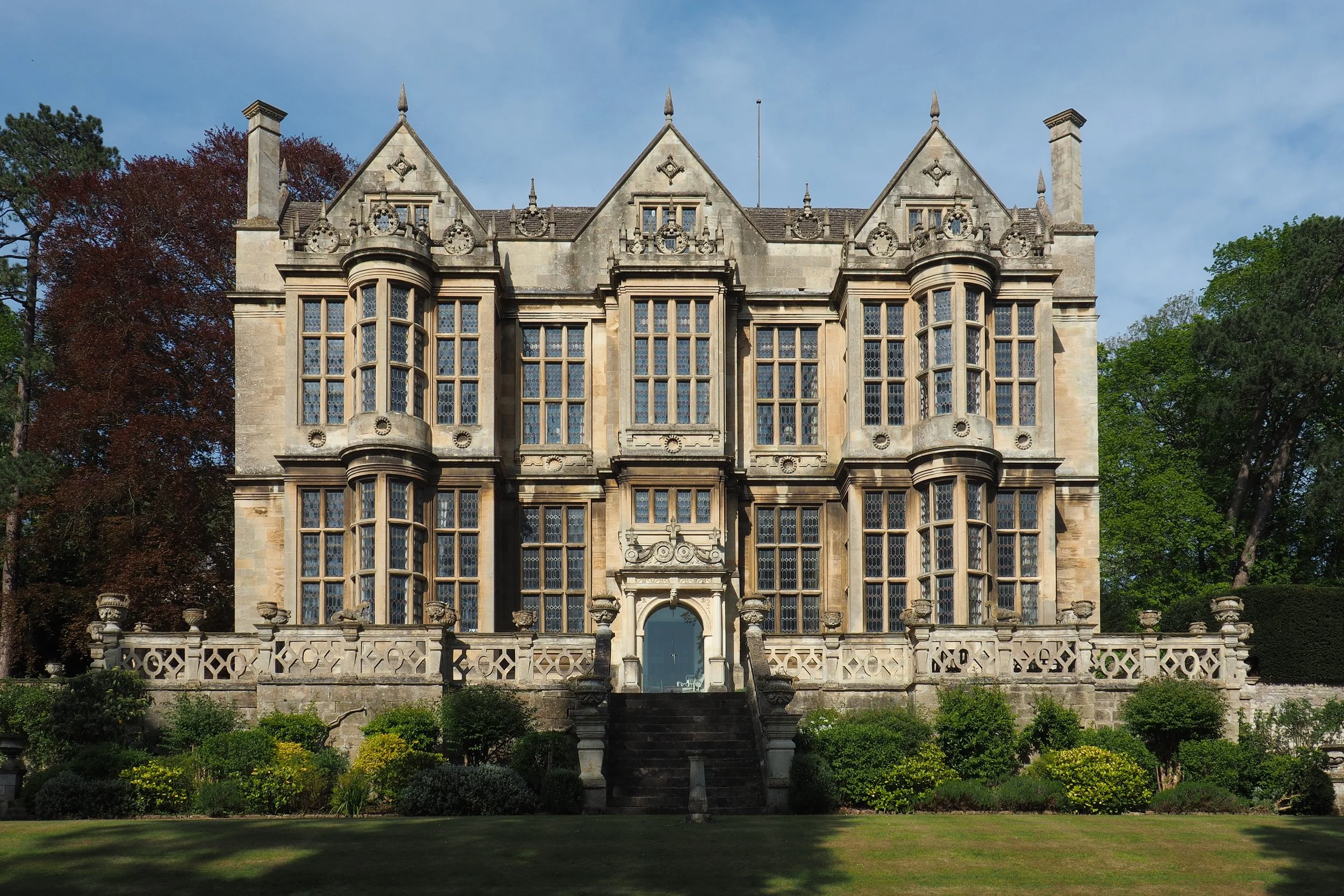 A large historic stone mansion with multiple tall windows and a decorative stone balcony, surrounded by a landscaped garden with bushes and trees, under a blue sky.