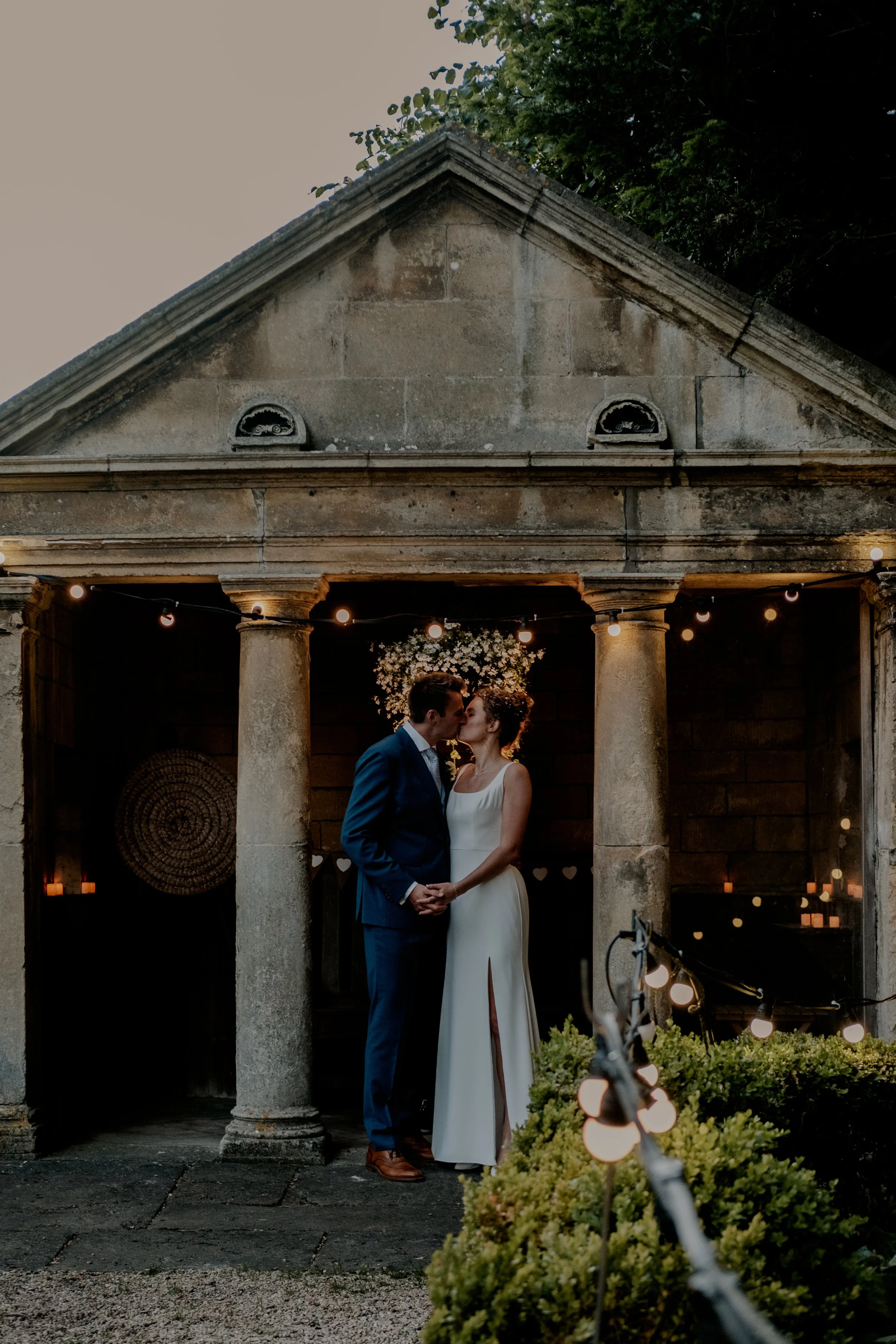 A bride and groom share a kiss under a stone portico with columns, string lights, and candles, during their wedding reception in an outdoor courtyard.