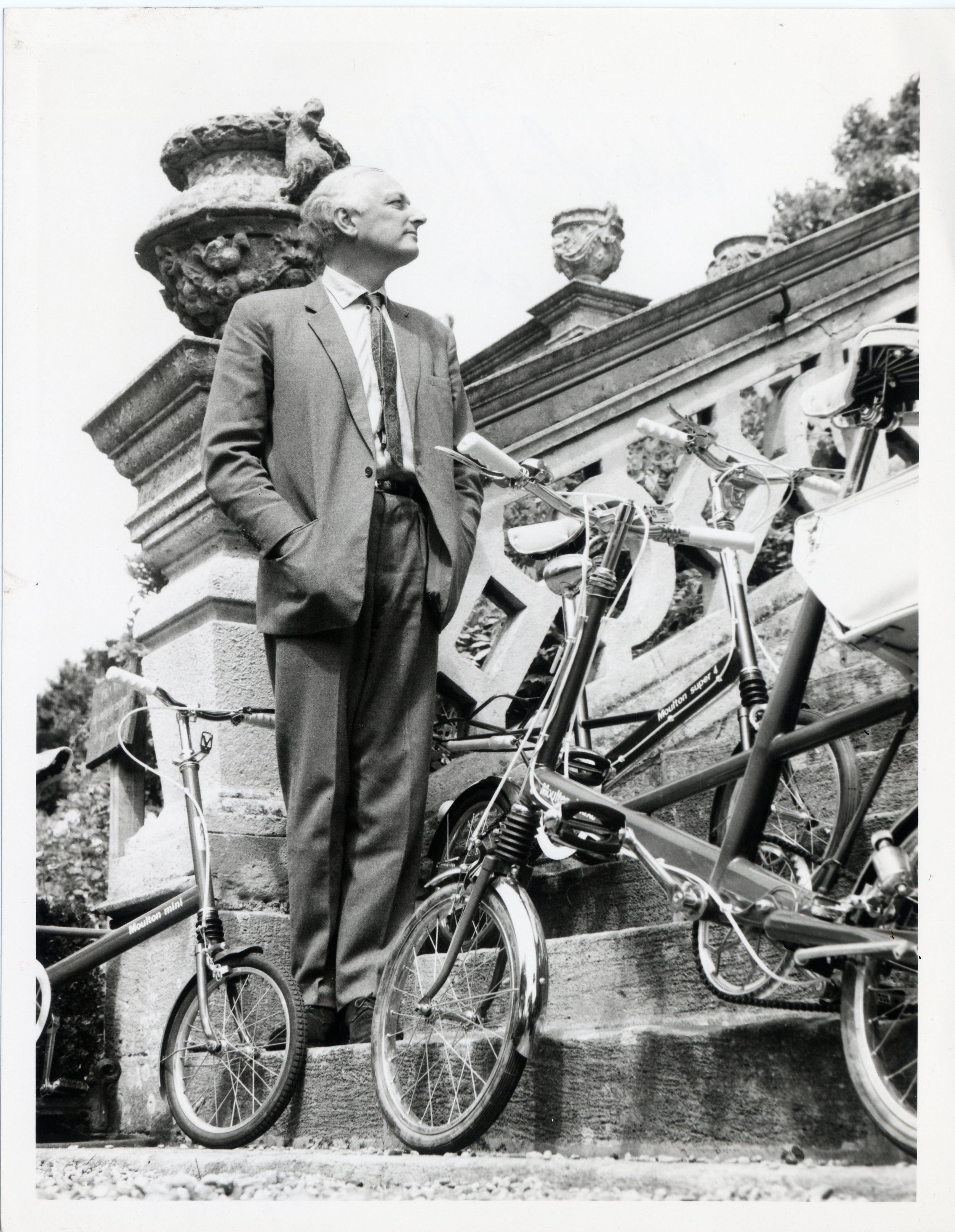 A man in a suit and tie standing next to several bicycles on steps with decorative stone balustrades and urns, looking upwards with a thoughtful expression.