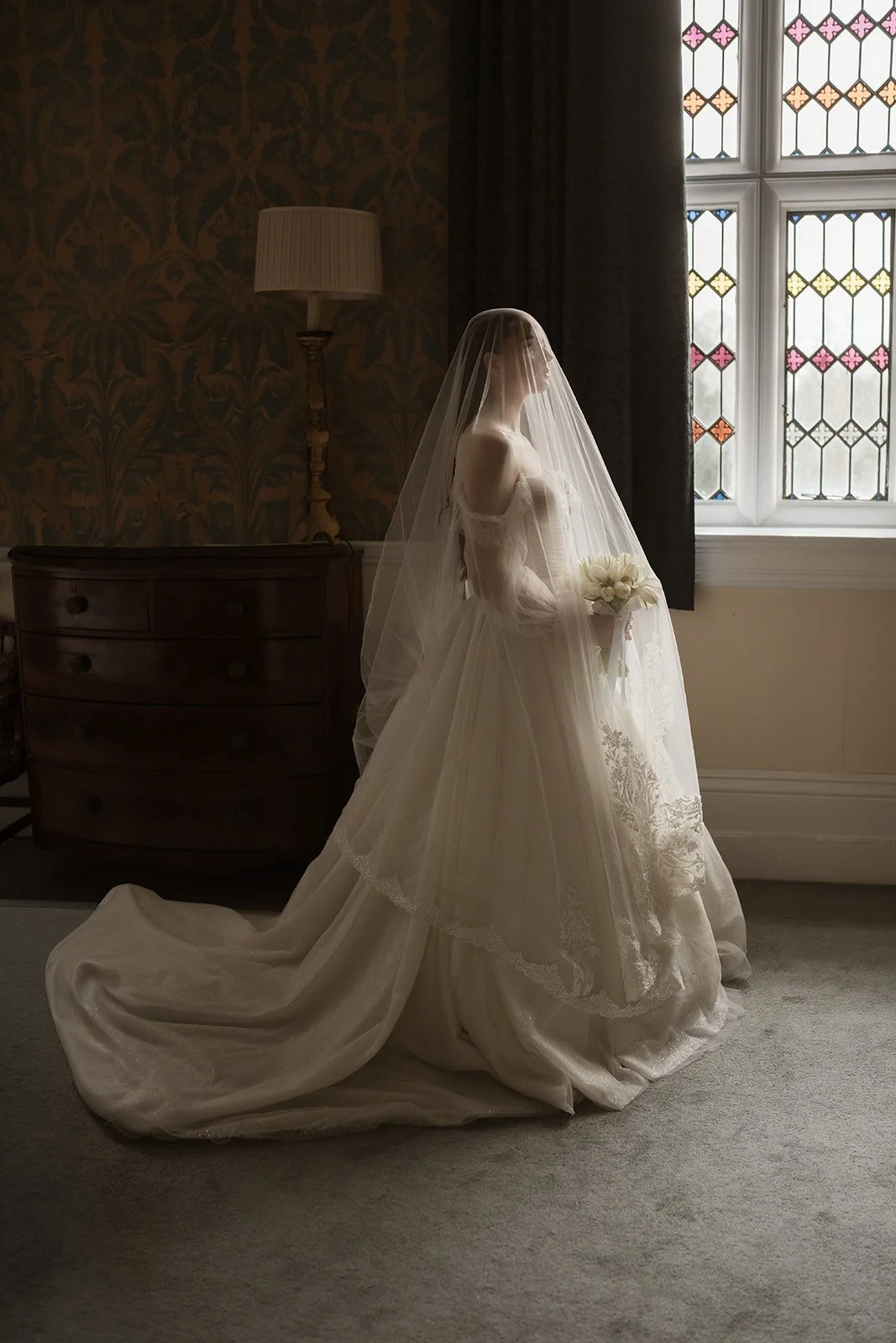 A bride stands by a stained glass window, wearing a long white wedding dress with a veil, holding a bouquet of white roses in a dimly lit room.