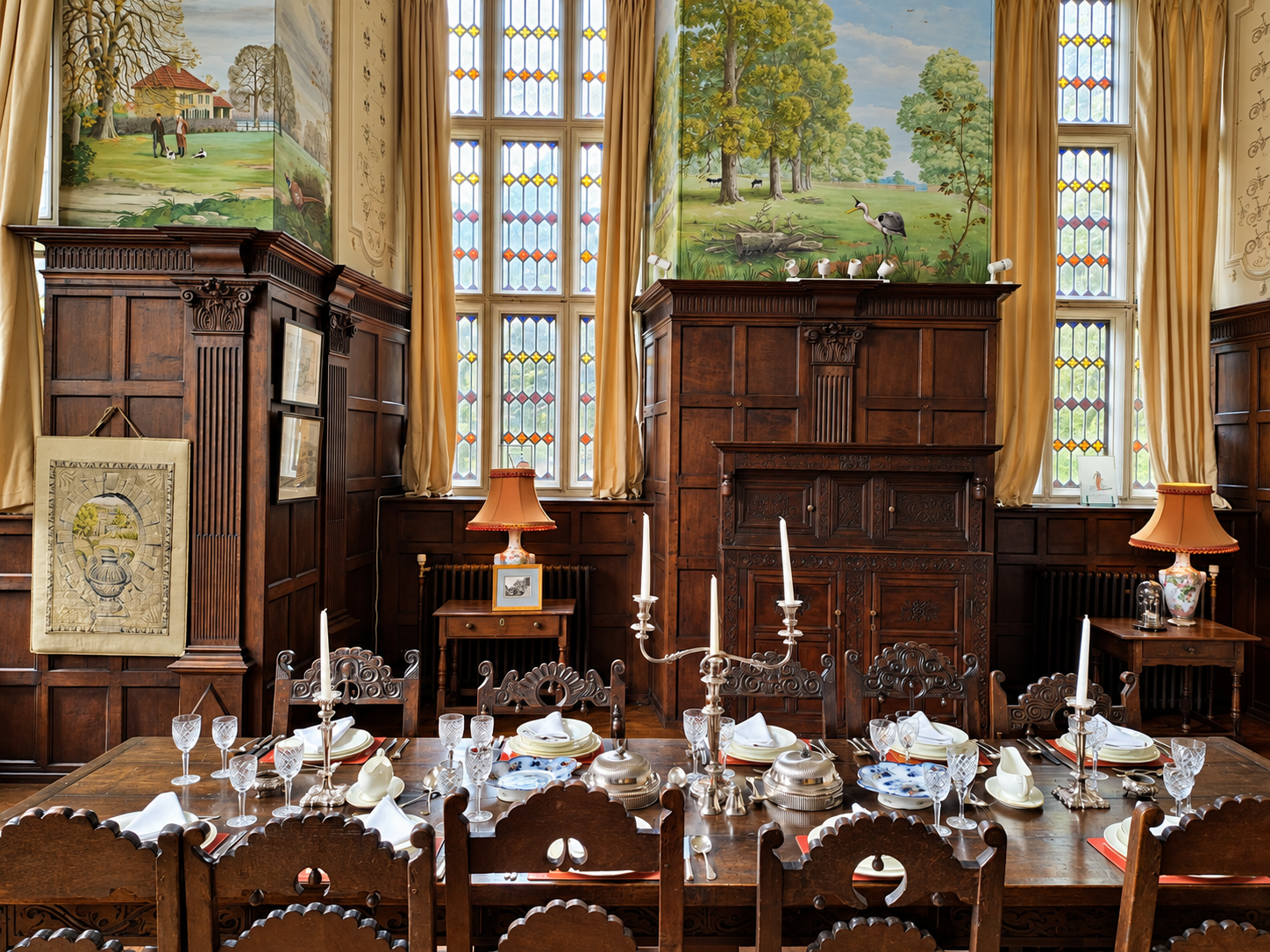 A dining room with a long wooden table set with white plates, glassware, silverware, candles, and tableware, surrounded by carved wooden chairs. The room features tall stained glass windows, dark wood paneling, and wall murals of nature scenes with t