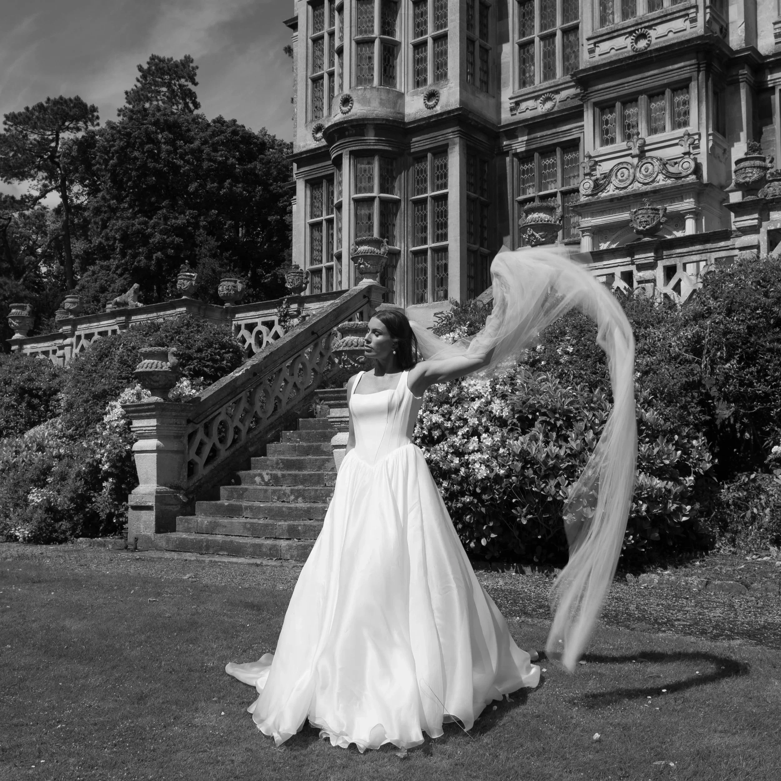 A woman in a flowing white dress standing on stone stairs in front of a historic building with ornate windows and decorative architectural elements. The woman is holding her veil as it blows in the wind, and there are trees and bushes surrounding the