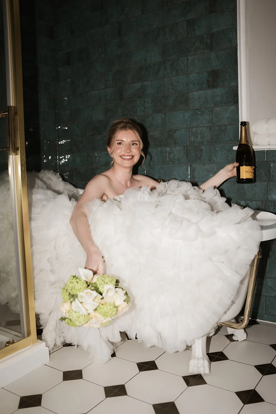 Woman in a white wedding dress sitting in a bathtub filled with white flowers, holding a bouquet in her left hand and a bottle of champagne in her right hand, smiling.