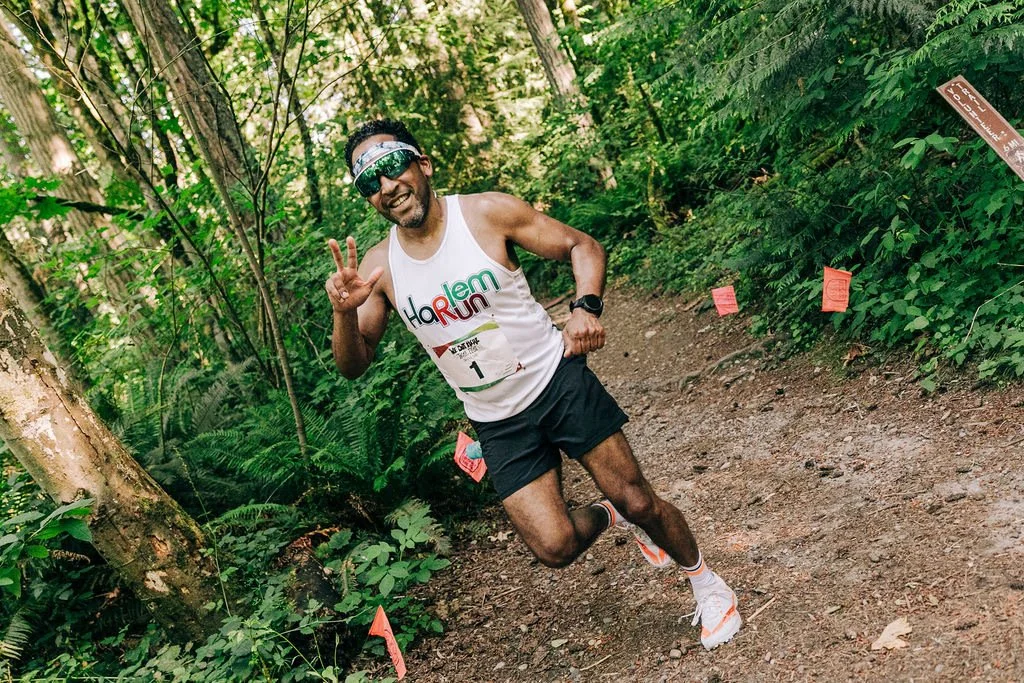 A joyfyl male runner making a peace sign with his right hand during a trail run in a lush green forest.
