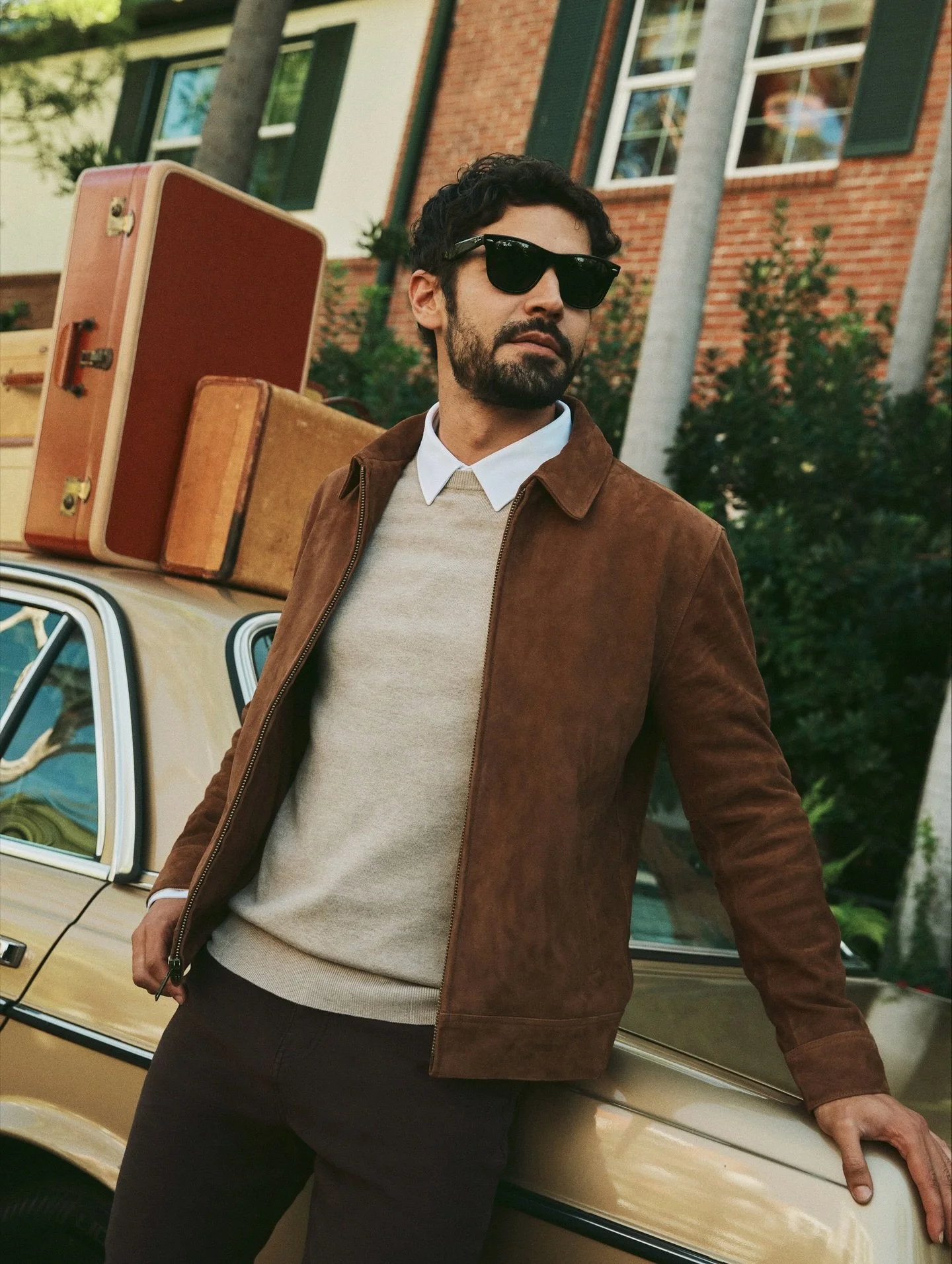 A man with dark curly hair and a beard wearing sunglasses, a 7 Diamonds brown suede jacket, a beige sweater, and a white collared shirt, leaning against a vintage yellow car loaded with suitcases on the roof, in front of a brick house.