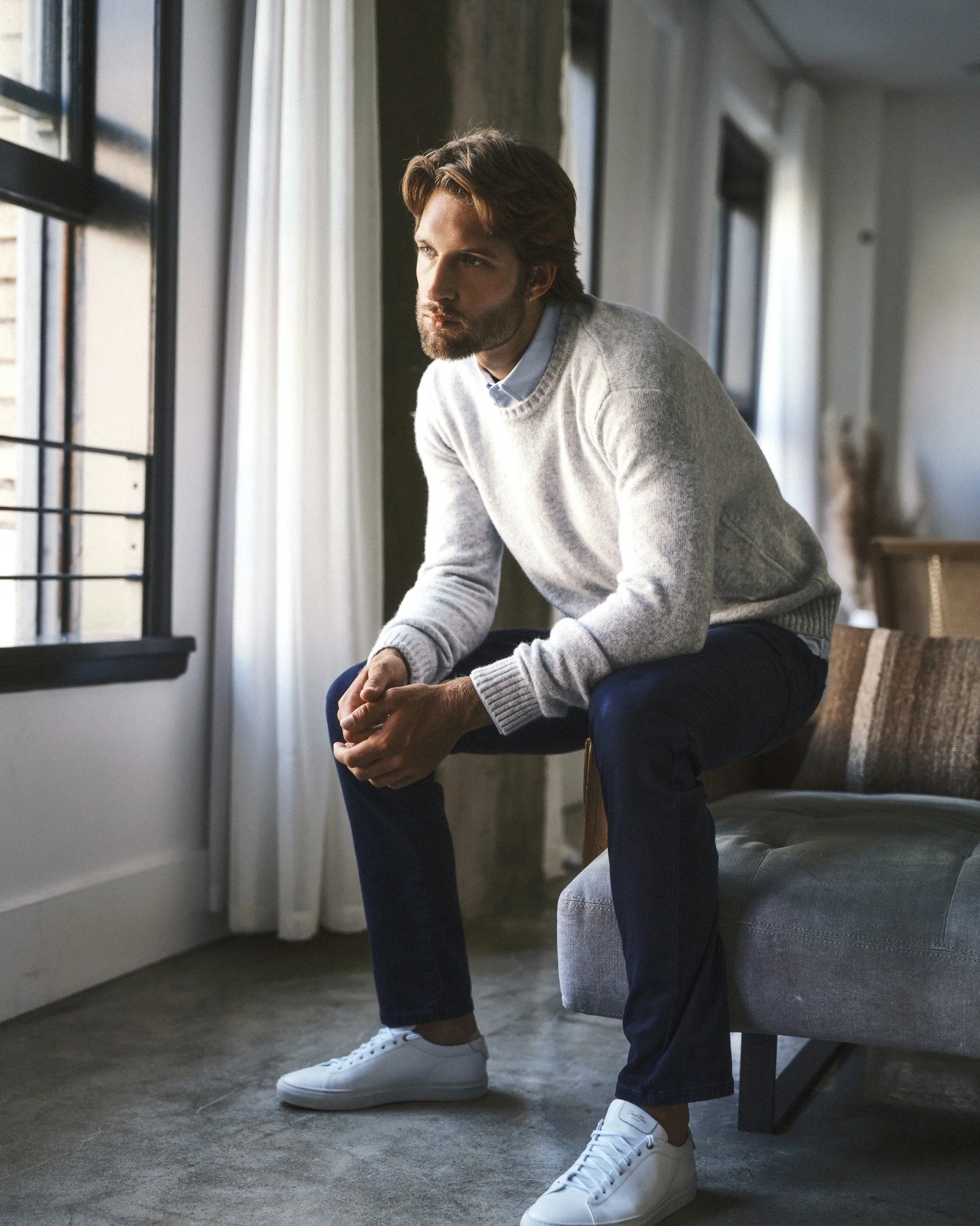 A man sitting on a sofa near a window, looking pensively outside. He is wearing a gray sweater, dark jeans, and white sneakers from 7 Diamonds.