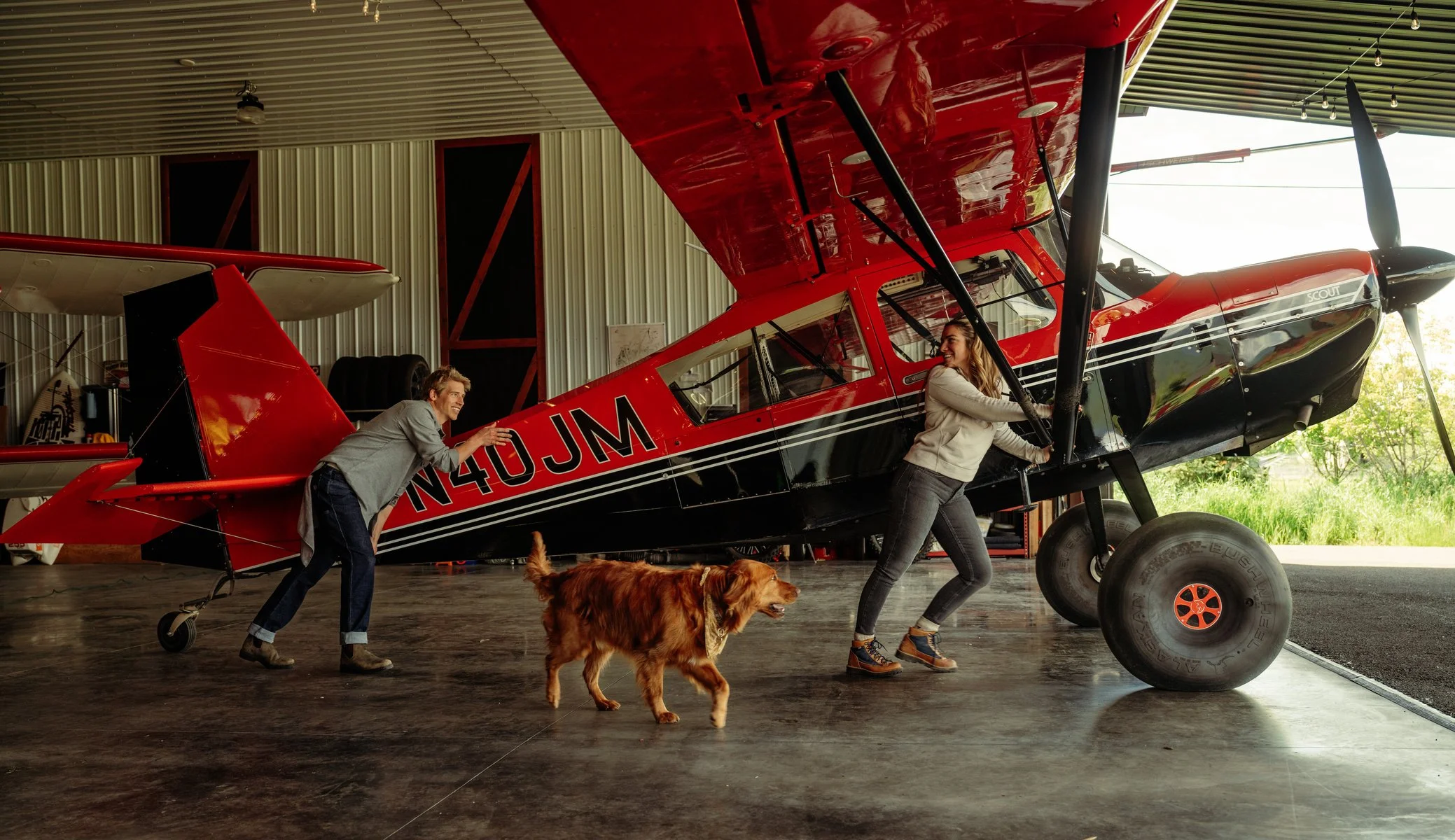 Two people and a dog pushing a red and black small airplane inside a hangar. The man and women are wearing Kuhl clothing.