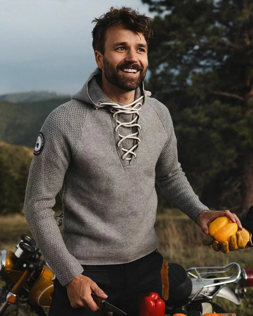 A smiling man outdoors with a motorcycle wearing a grey laced hooded Amundsen sweater, surrounded by trees.