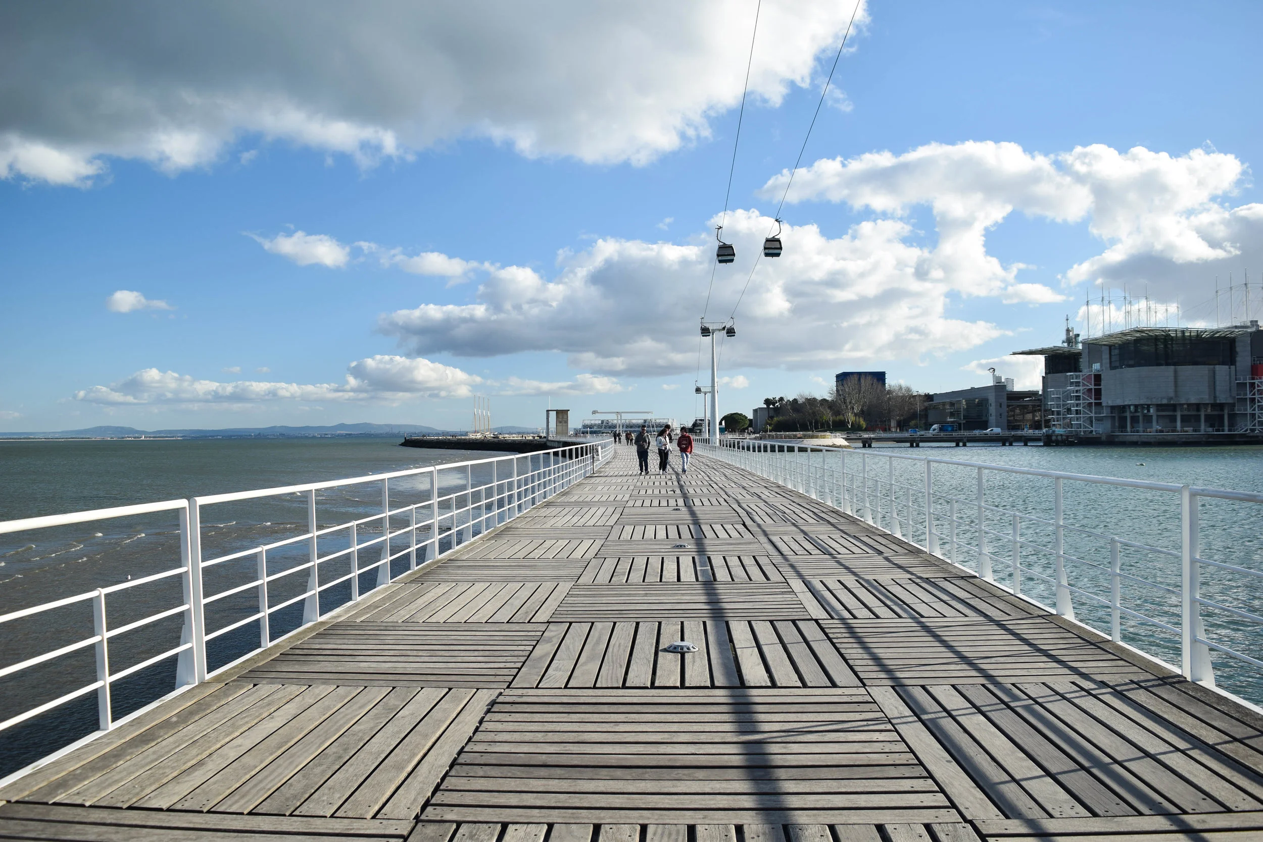 A wooden pier with a white railing extending over water, with a few people walking, a sky with clouds and clear blue, and modern buildings in the background.
