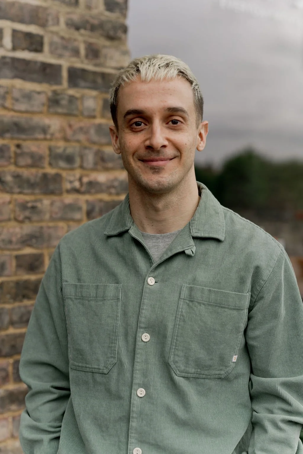 A man with light blonde hair and a light green corduroy shirt standing outdoors against a brick wall, with cloudy skies in the background.