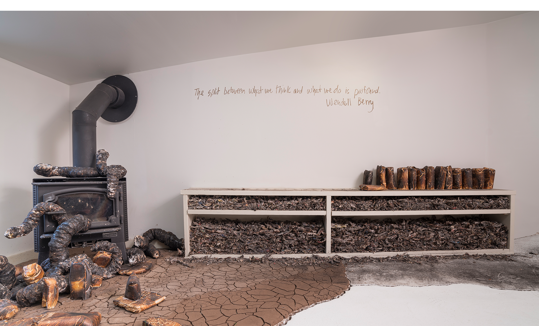 Installation detail with wood burning stove, charred newspaper fragments rammed into bookshelf, and cast plaster organic forms with charred consumer goods casts, cracked earth and ash. Wall texts drawn in Kansas mud, Wendell Berry.