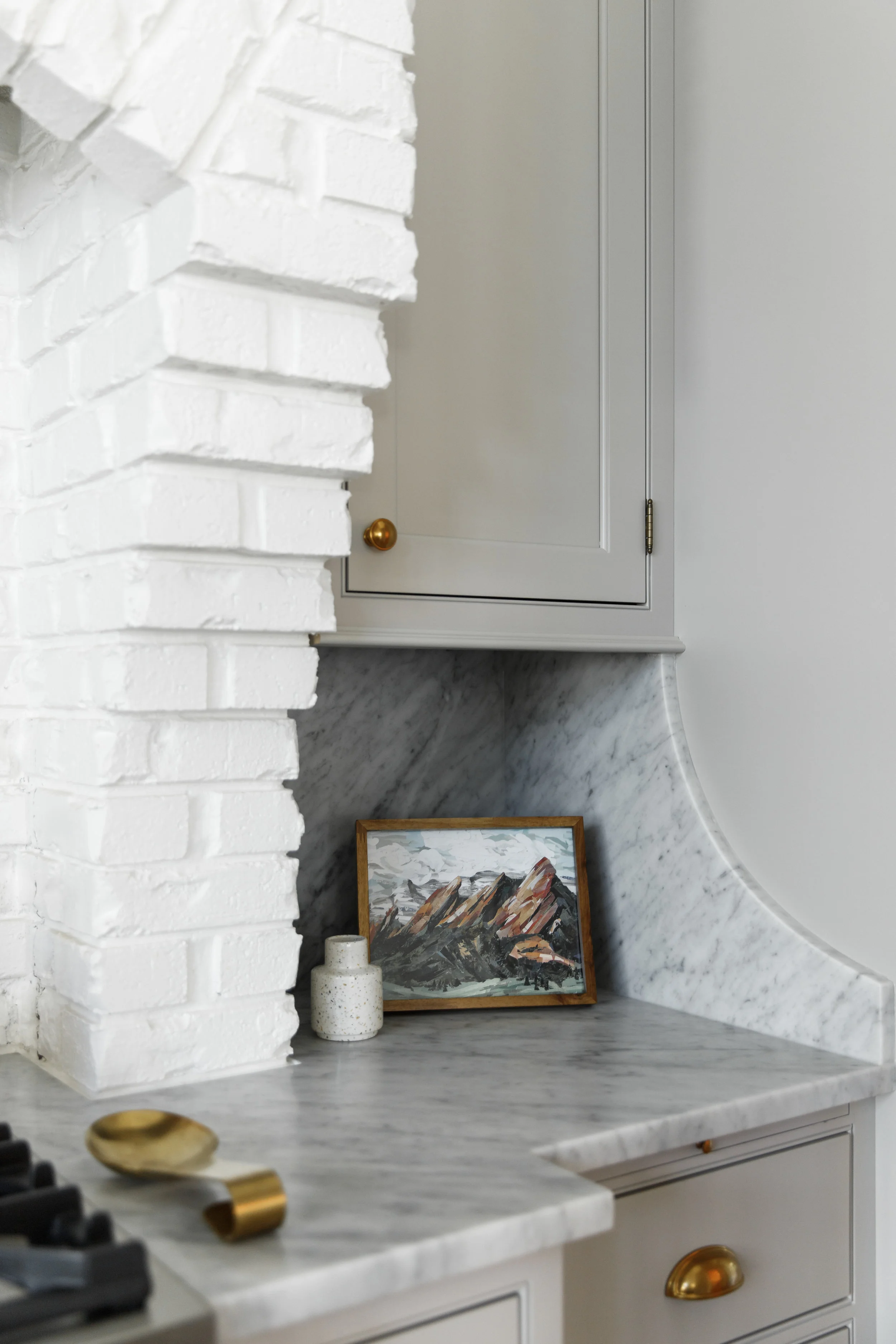 Light gray cabinets with gold hardware, picture frame, brick range hood and full height honed Carrara Marble countertops with backsplash.