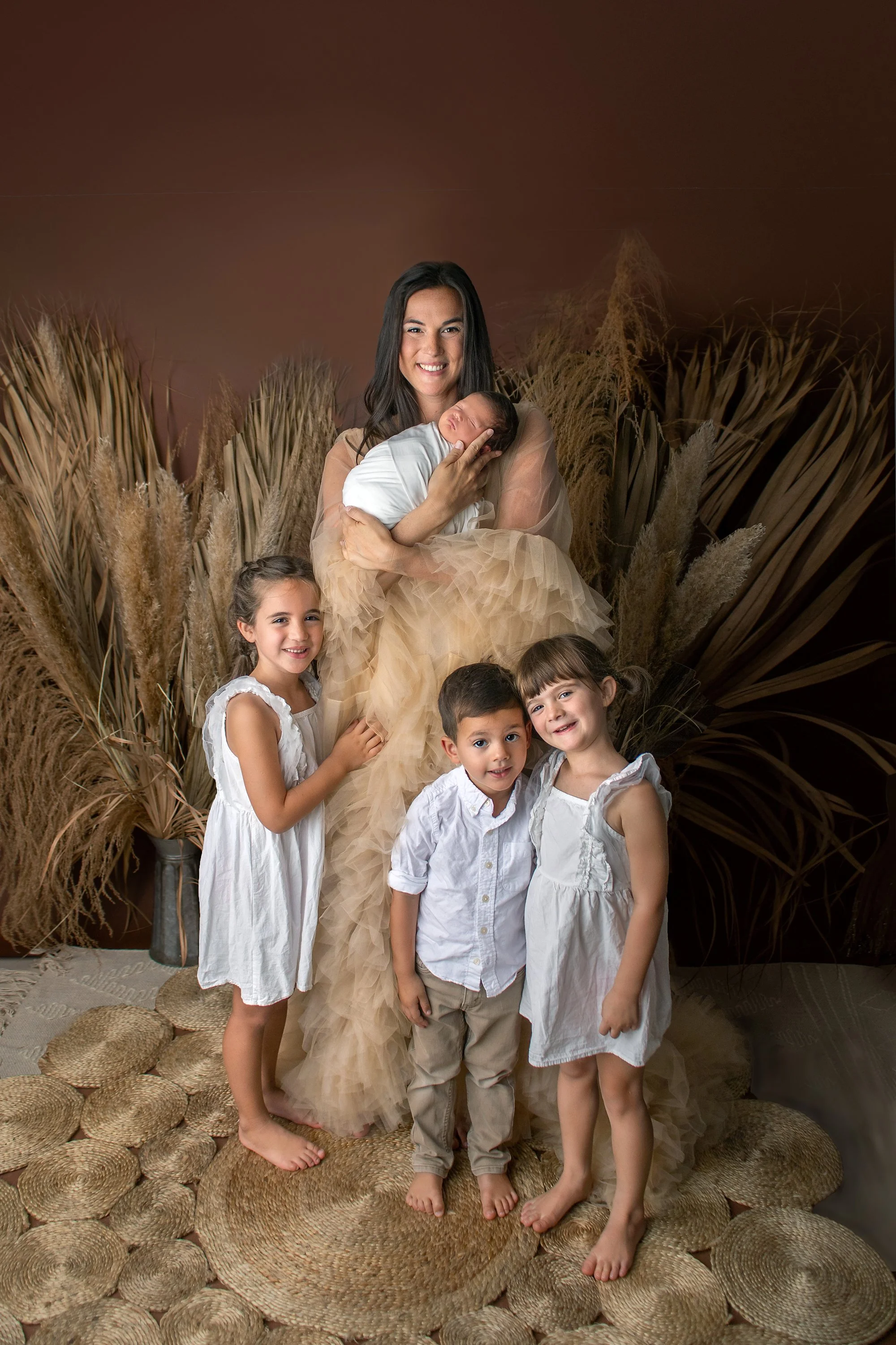 Baby boy in white wrap with siblings in white and mom in tulle dress gown with boho palm fronds and grasses and rug