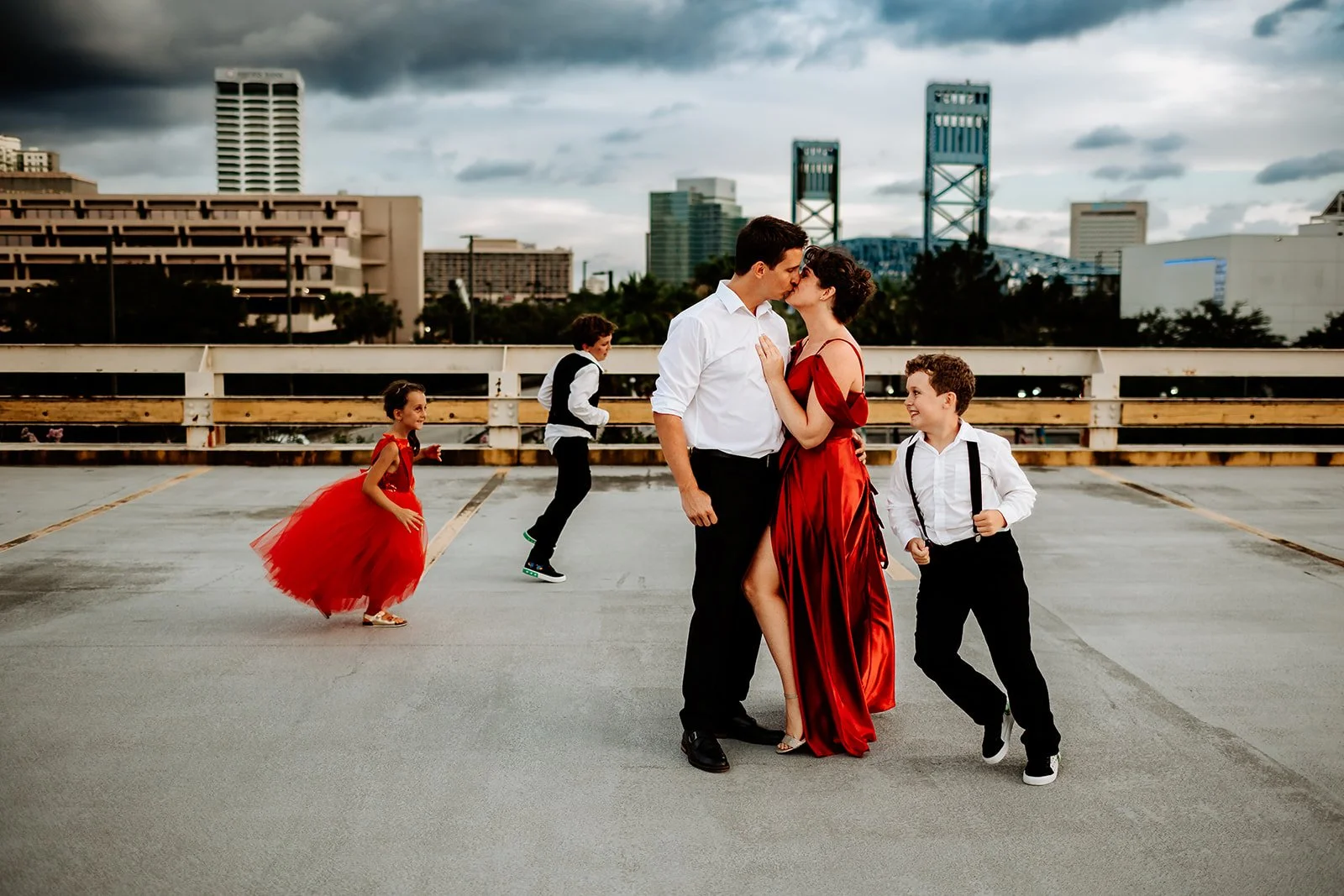 JAX FL family on rooftop in elegant attire, parents kissing, children playing, urban skyline backdrop.