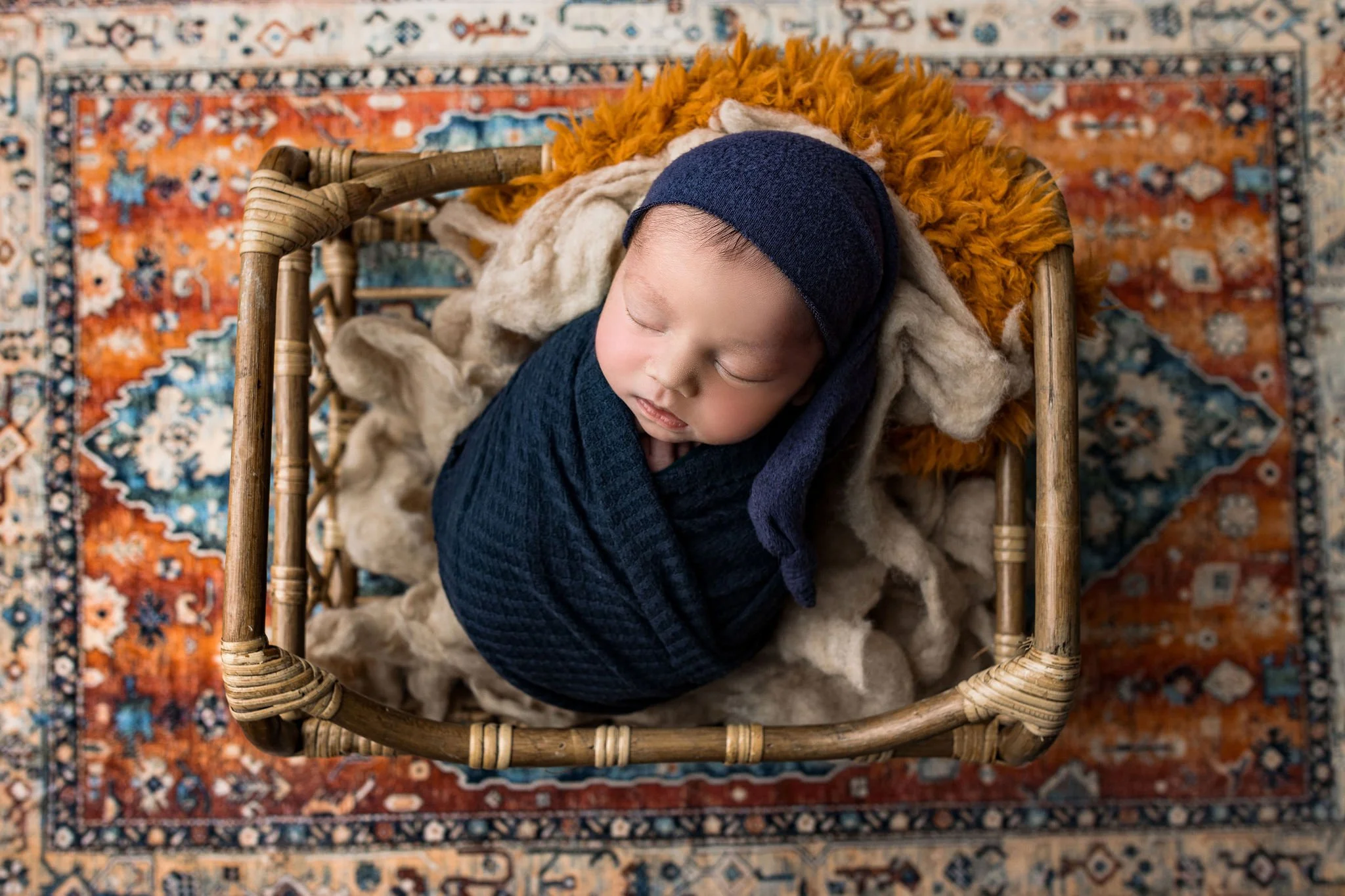 Exclusive newborn photographer in St. Augustine FL with baby boy wearing navy wrap in bucket with rust tone rug in portrait studio