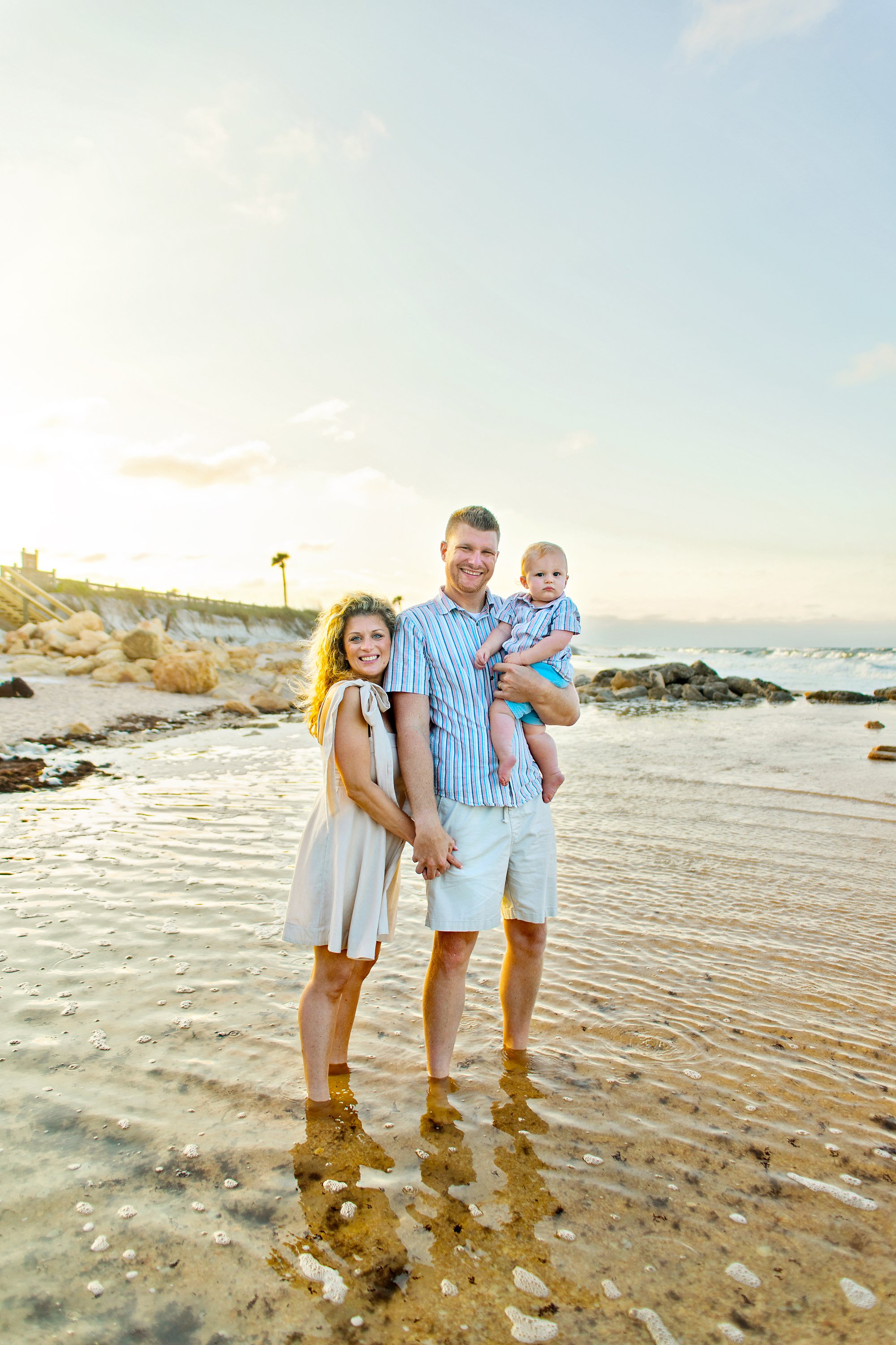 JAX Beach, FL family standing on a beach with a baby in father's arms, sunlight and sand in the background. Photos by Jacksonville's number one family photographer.