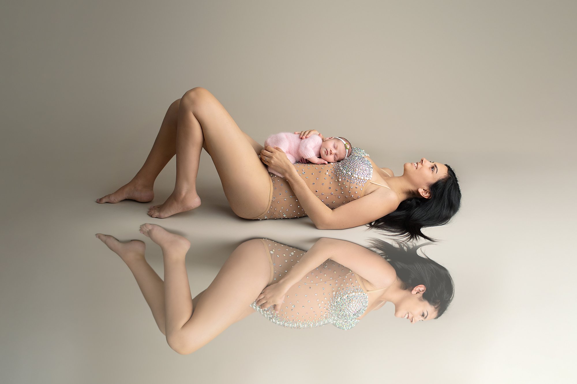 A woman in Ponte Vedra Beach, FL in a sparkling bodysuit lies on the floor with a sleeping baby in pink, her reflection visible. Best photography studio in PVB, florida