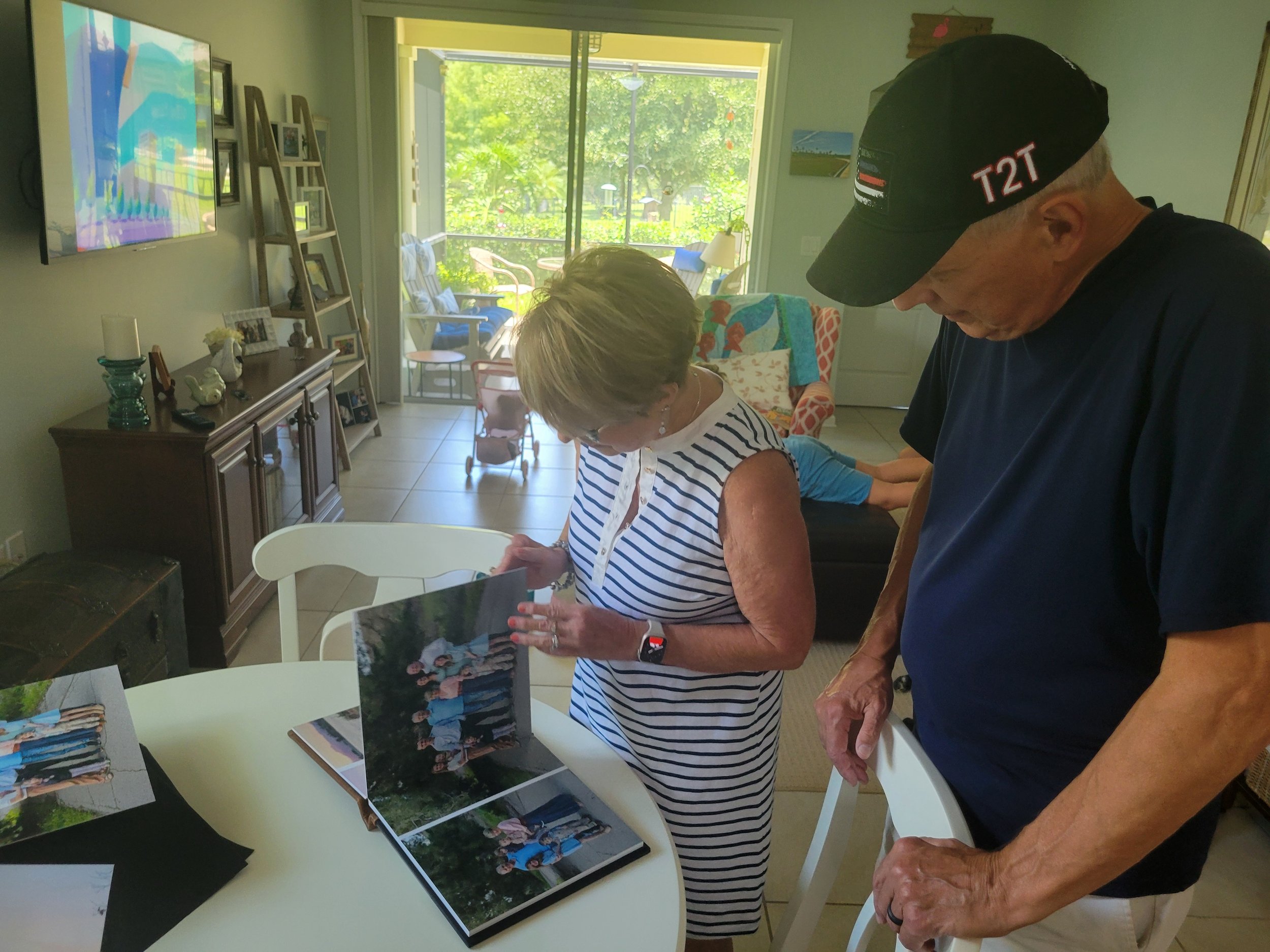 Grandparents looking at photos from extended family photo shoot while on vacation. Photo album has pictures at the beach with kids during sunset with greenery and park in background.