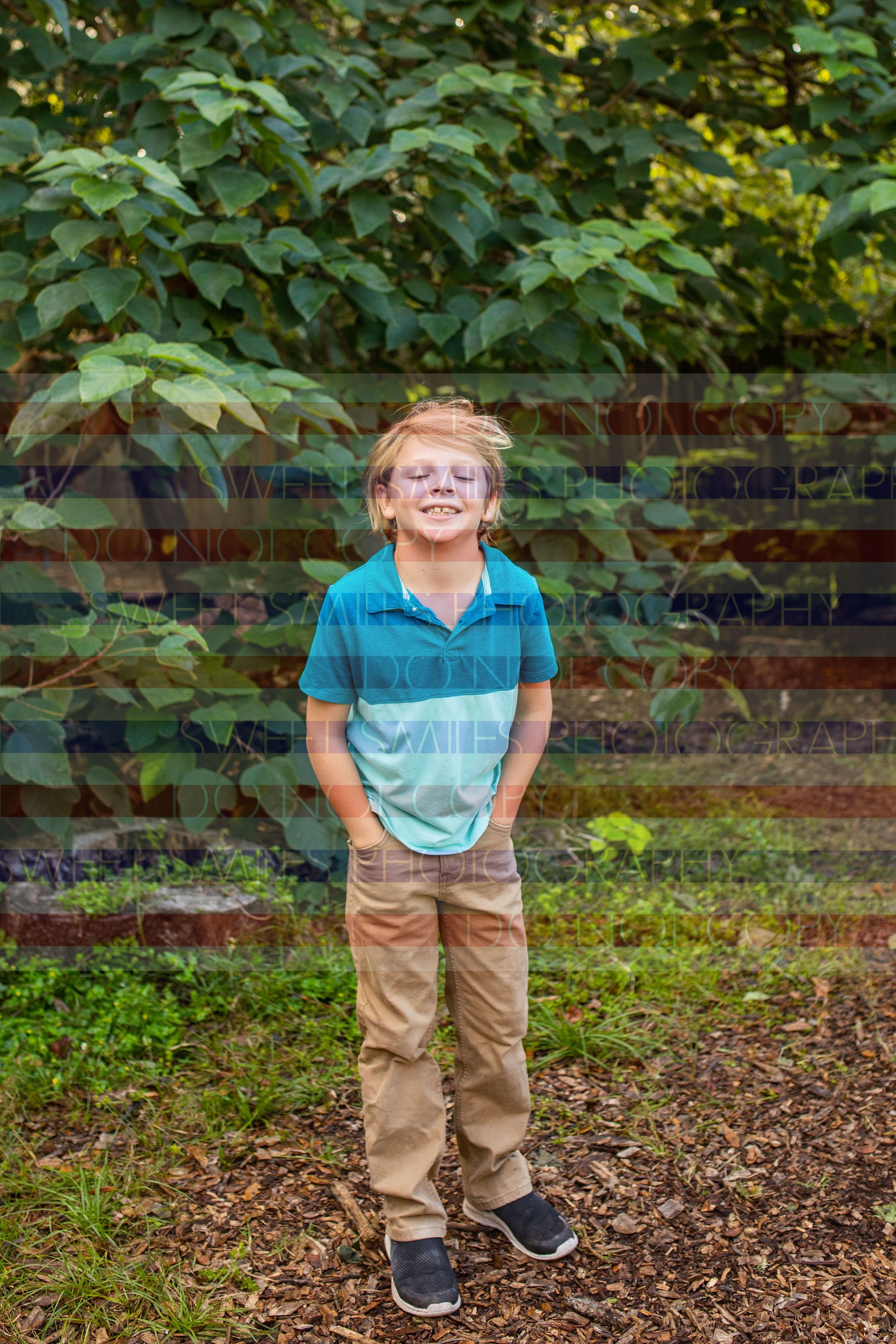 A smiling child stands outdoors in front of green foliage, wearing a blue shirt, khaki pants, and black shoes for family photo session in ST. Augustine, FL. 32086