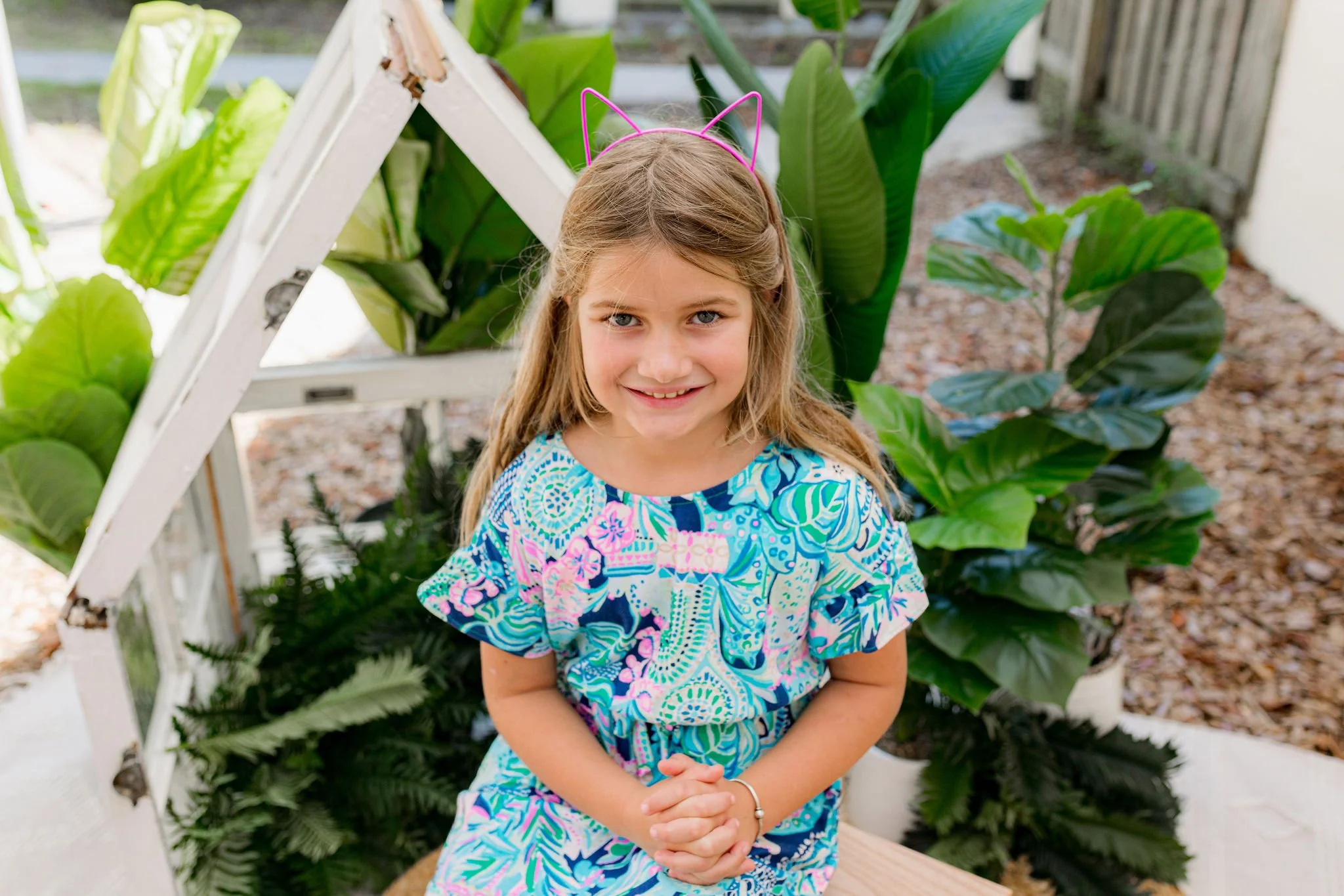 Young girl in Jacksonville, FL during photo session wearing blue floral dress and pink cat ear headband, sitting outside with plants and wood chips in background.