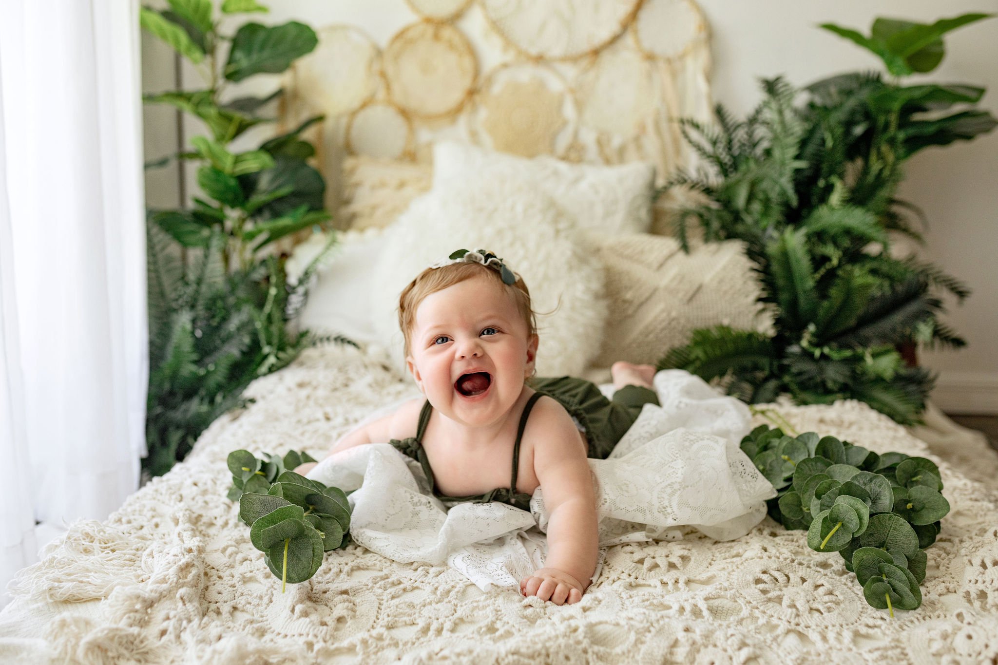 Best Jacksonville family photographer in FL with private studio doing 6 month old baby pictures with boho and greenery as the set up and baby in dark green clothes laying on bed with vintage lace blanket