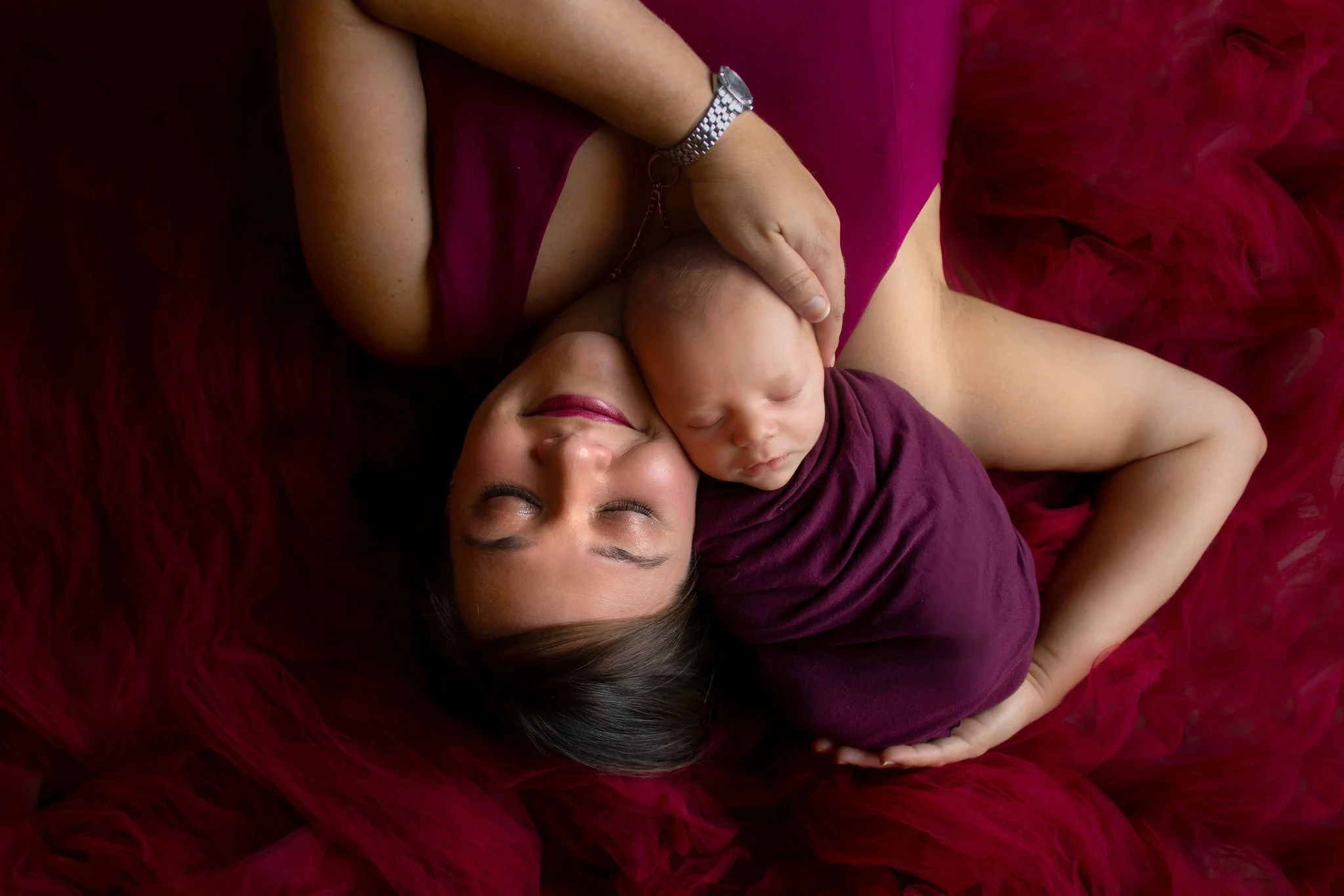 Jacksonville FL mother lying on a burgundy background, cradling a sleeping baby wrapped in a matching cloth in best photography studio.