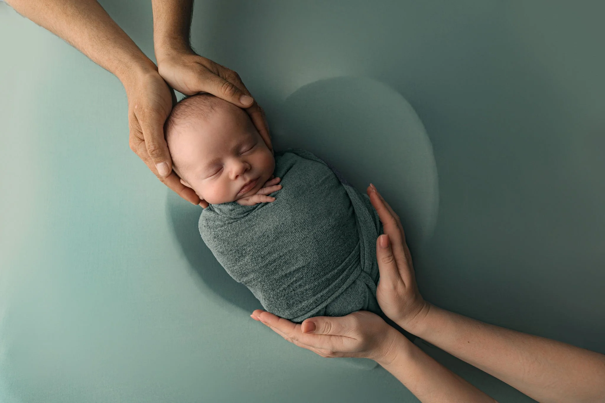 Sage green wrap and background with parents hands around hearth bowl with newborn baby in photo session at St. Augustine, FL studio