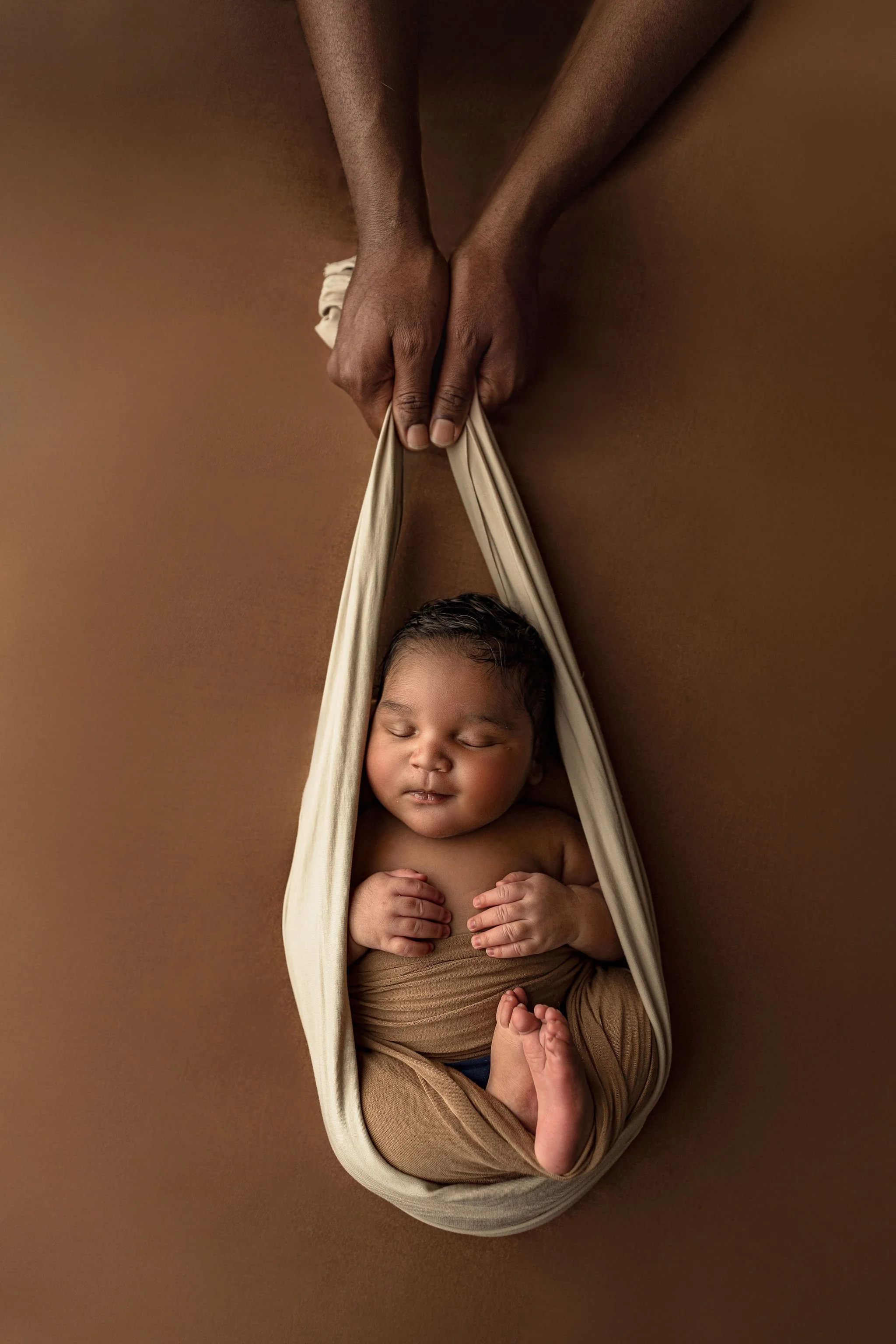 Newborn photographer in St. Augustine, FL with african american baby boy on earthy brown background with father's hands around wrap