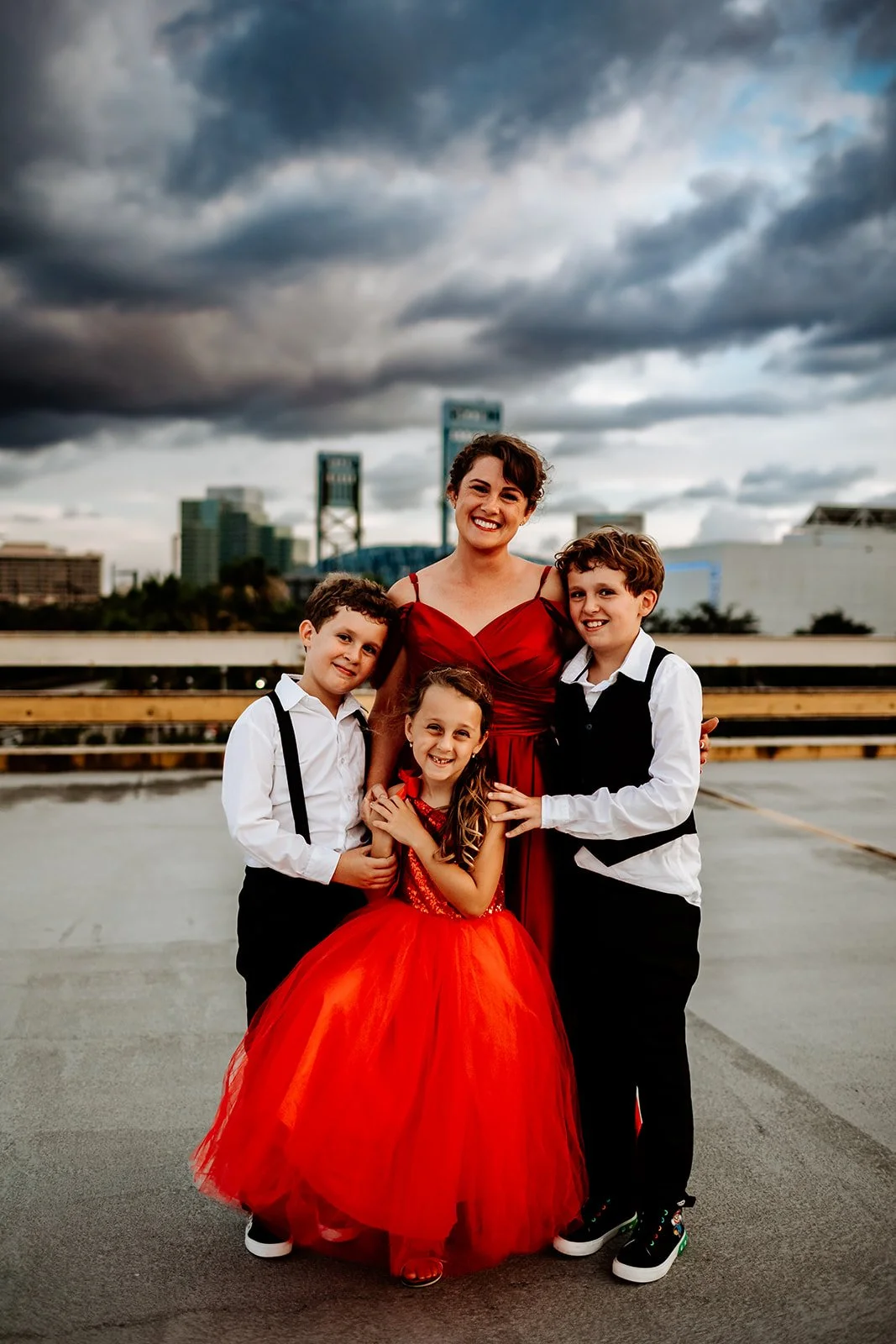 A group of four people in Jacksonville FL, three children and one adult, dressed in formal red and white attire, standing outdoors with a cloudy sky and city buildings in the background.