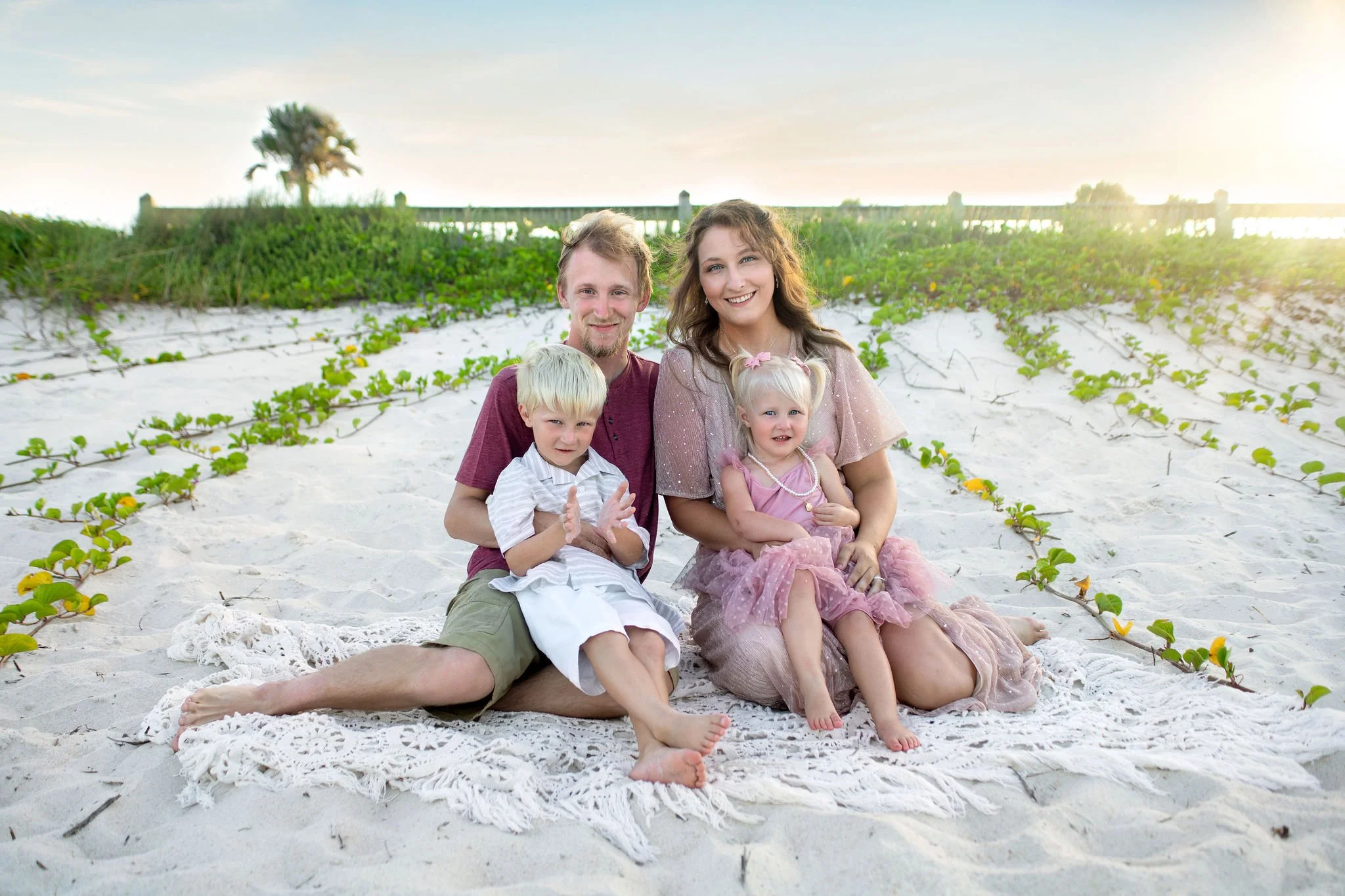 Family on vacation in St. Augustine, FL sitting on beach blanket at sunset with greenery in background.