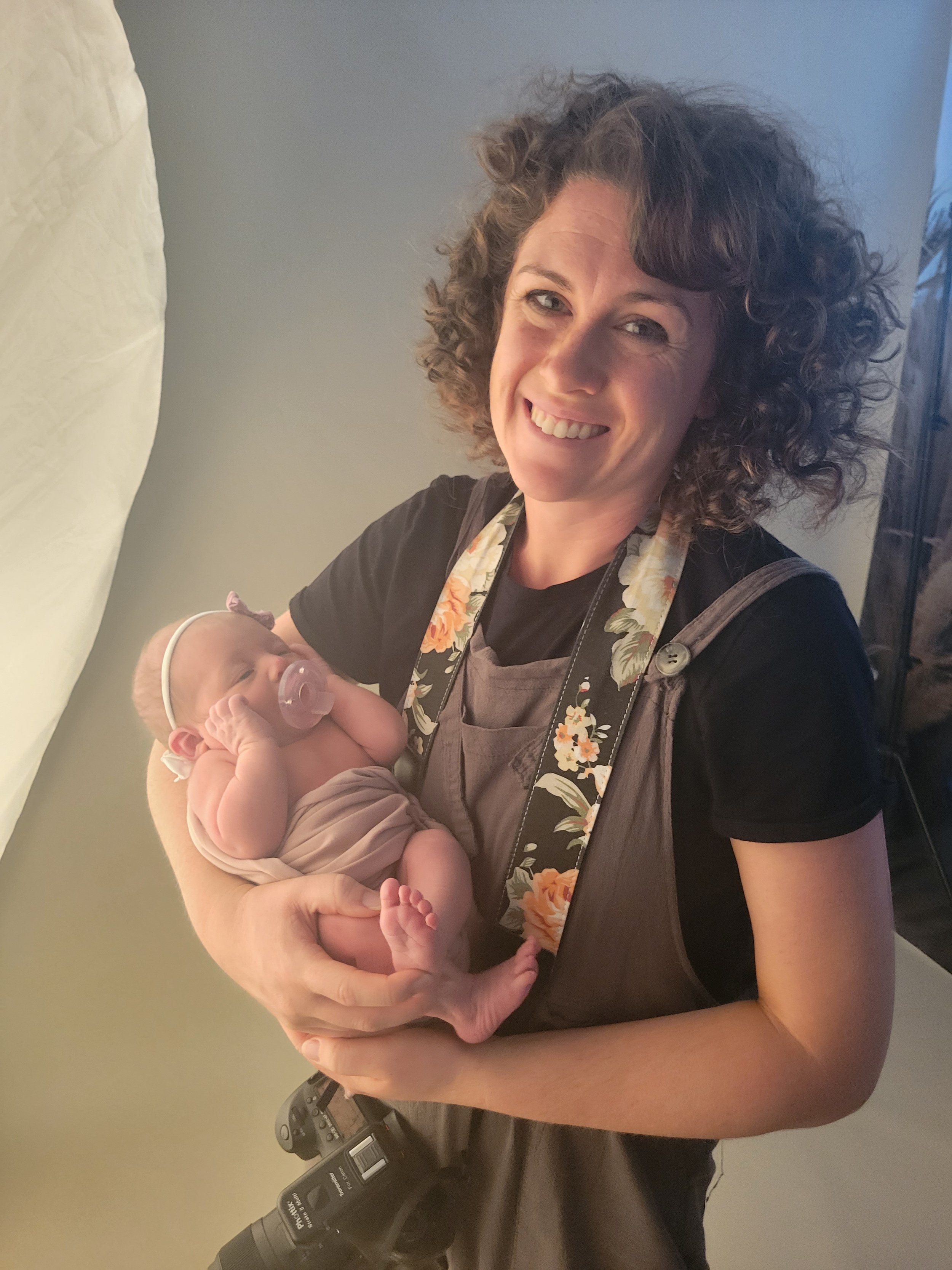 World Golf Village, FL newborn photographer holding a swaddled baby with a pacifier, smiling at the camera, camera strap around her neck.