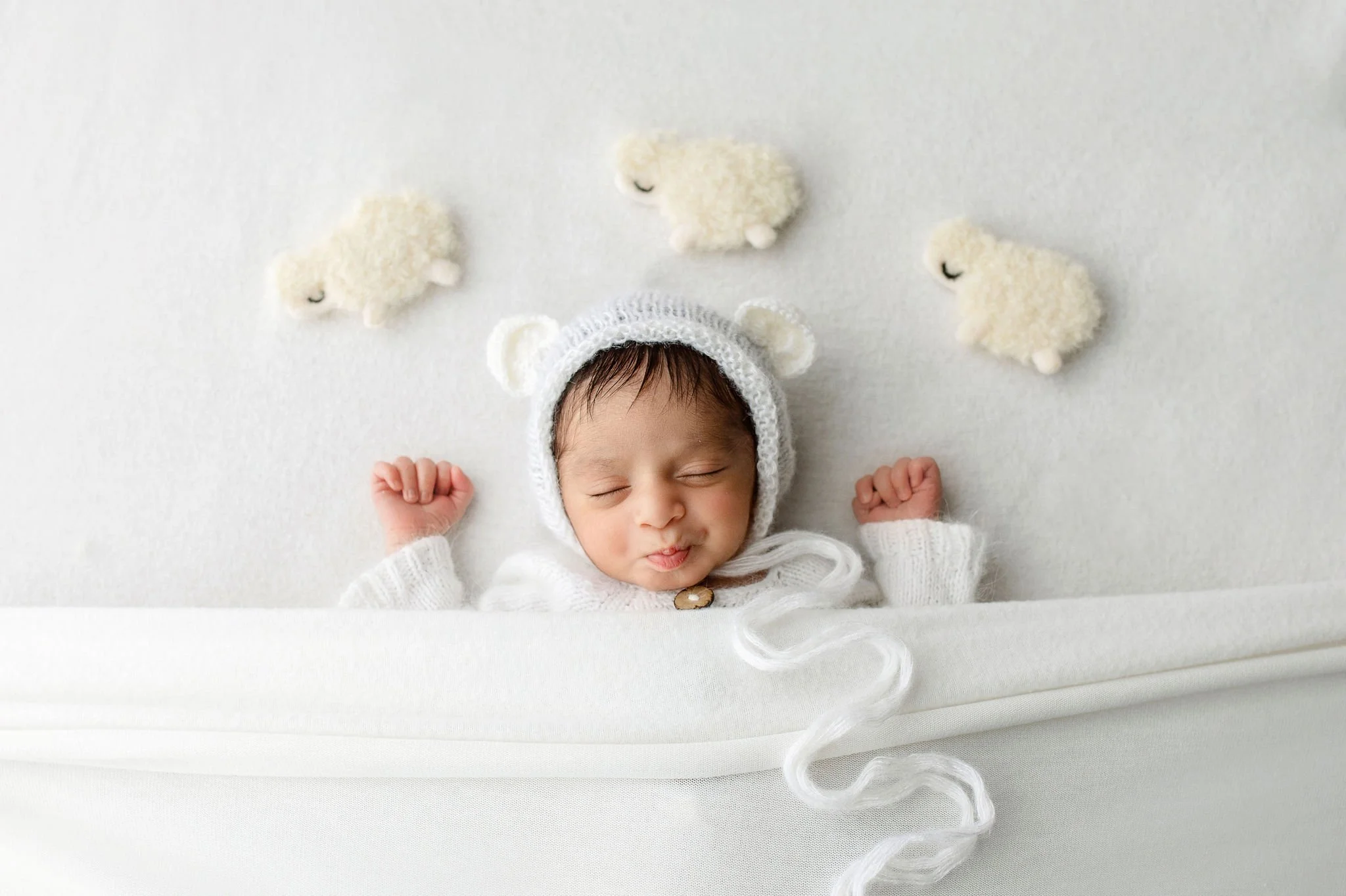 Indian newborn photo shoot in St. Augustine FL with sleeping baby in white outfit with bear ears, surrounded by plush sheep toys.