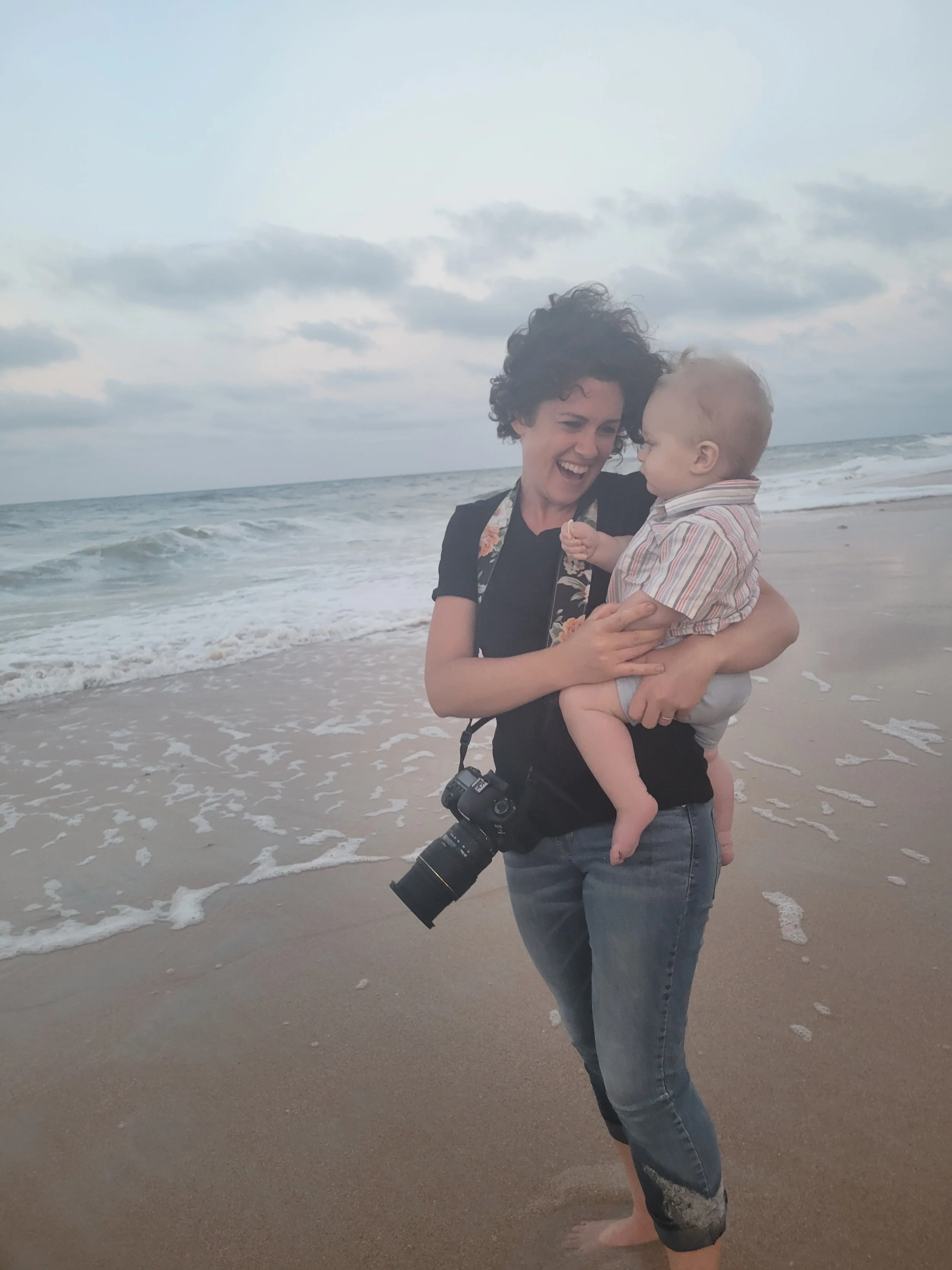 Fine art photographer in Ponte Vedra, FL holding baby on a beach with waves in the background, camera hanging from her shoulder.