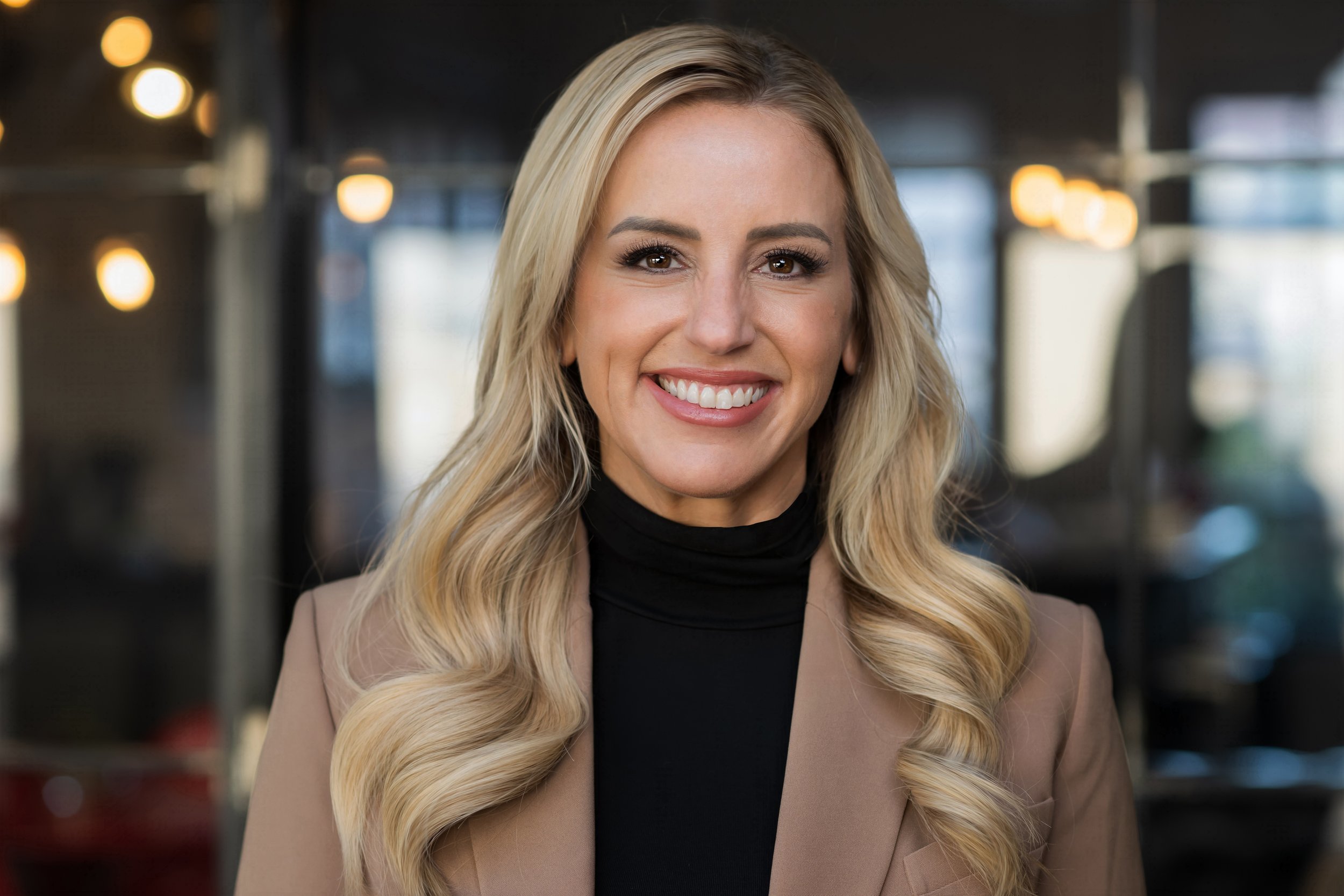 Smiling woman with long blonde hair wearing a brown blazer and black top, standing in an office setting.